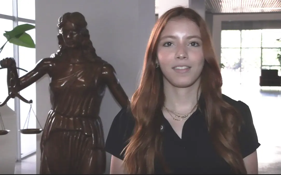 A imagem mostra a acadêmica Brenda. Ela é uma jovem de pele clara, olhos escuros e cabelos lisos e longos, ruivos. Ela veste uma camisa preta, de mangas curtas. Atrás dela, aparece a estátua de Artêmis, a deusa da Justiça