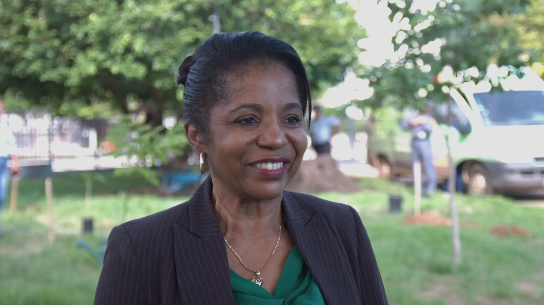 foto horizontal colorida da presidente do TRT, desembargadora Adenir Carruesco. Ela é uma mulher negra e está com cabelos presos. Usa uma blusa verde e blaser preto. ao fundo está o local onde foi inaugurado o bosque