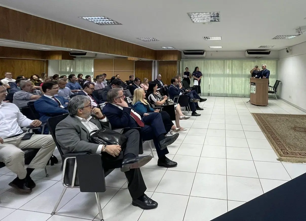 fotografia colorida de sala de aula. Homem está atrás de púlpito e fala ao microfone. À frente dele, pessoas sentadas à cadeira prestam atenção ao que é falado.