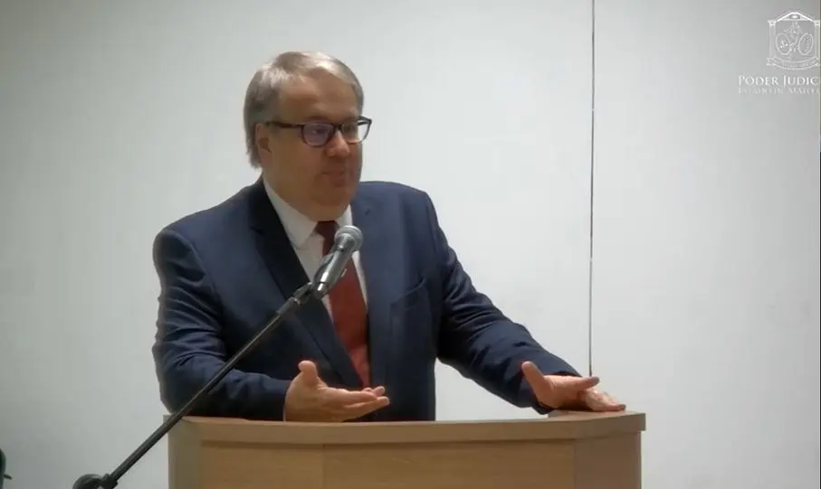 Homem veste terno escuro, gravata vermelha e blusa branca. Ele estava em pé atrás do púlpito e fala ao microfone.