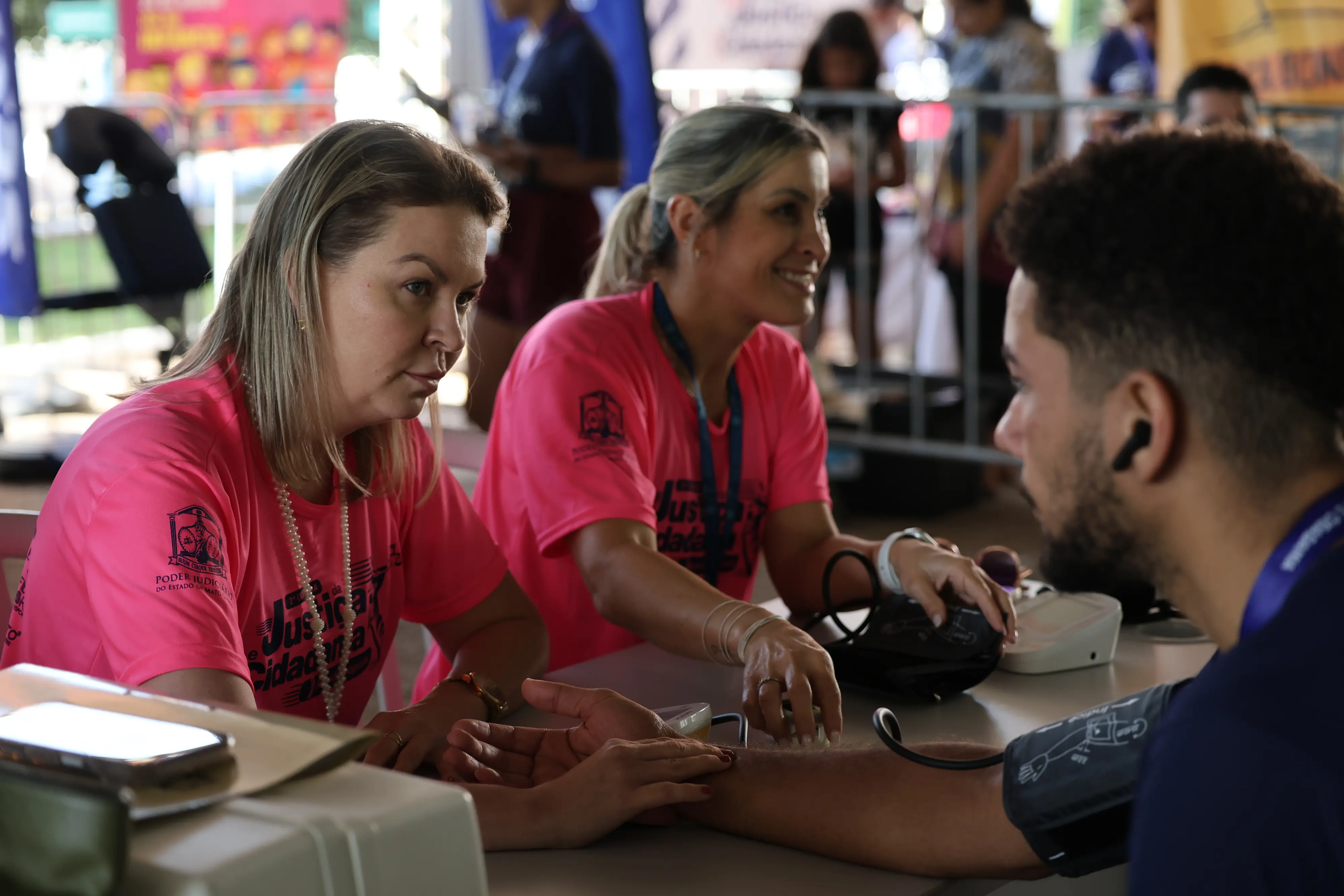 Duas mulheres vestidas de rosa auxiliam um homem durante aferição de pressão arterial em estande de saúde montado para o evento esportivo.