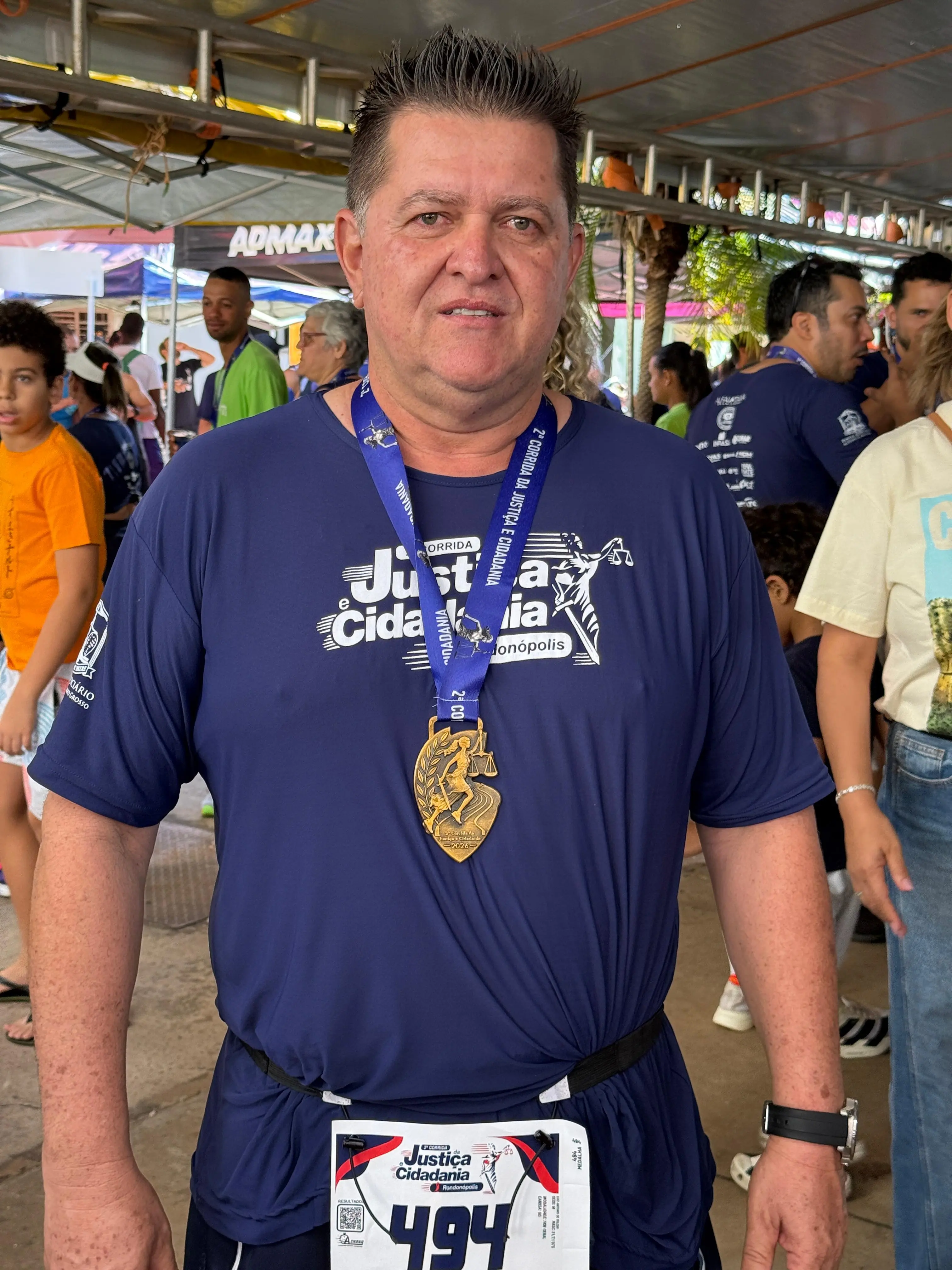 Um homem exibe medalha de participação pendurada no pescoço, com número de peito ainda afixado à roupa. Veste camiseta azul-marinho alusiva à corrida, em ambiente coberto.