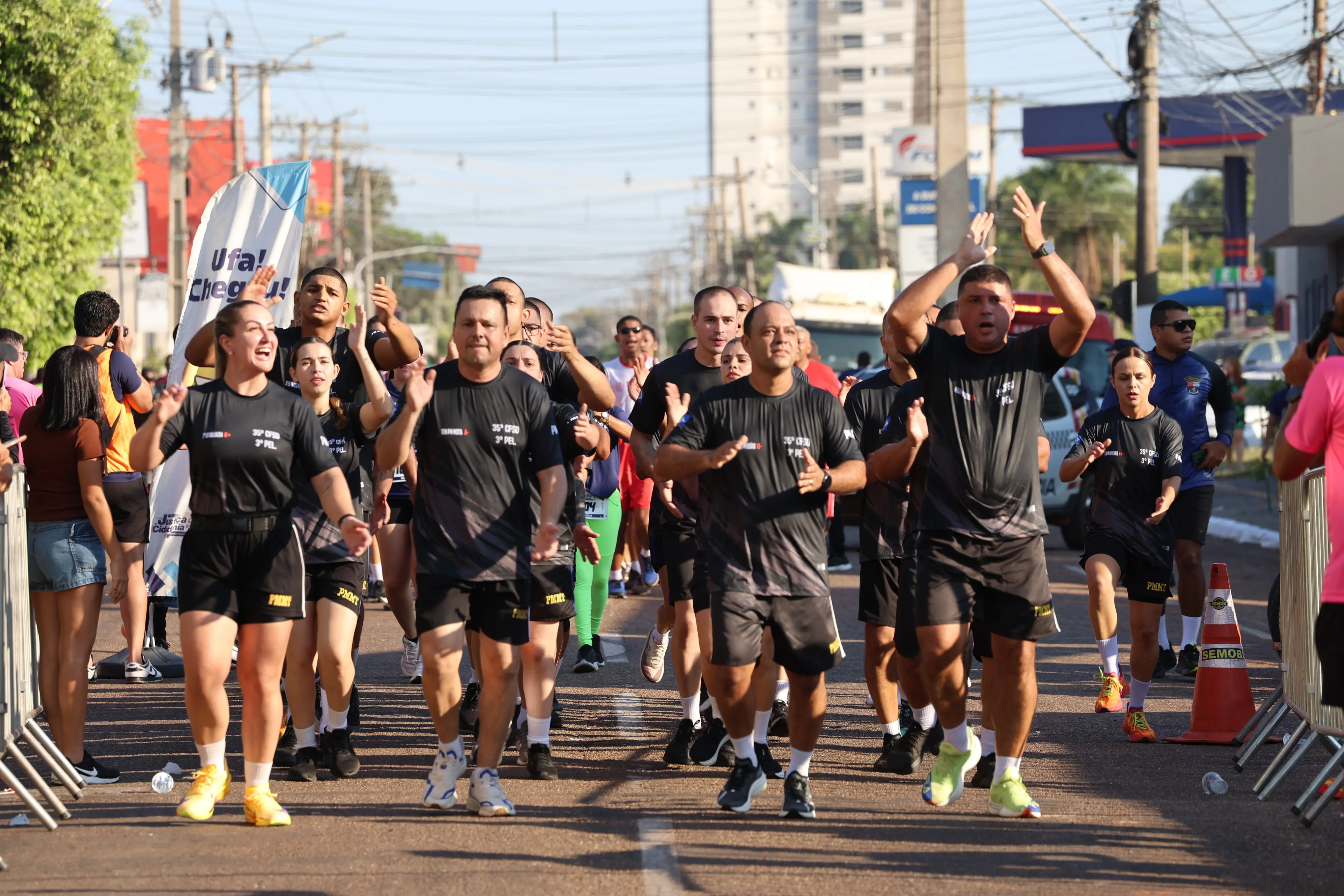 Grupo de policiais uniformizados corre em via pública, com as mãos erguidas em comemoração. Trajam camisetas pretas com identificação de corporação durante prova esportiva.
