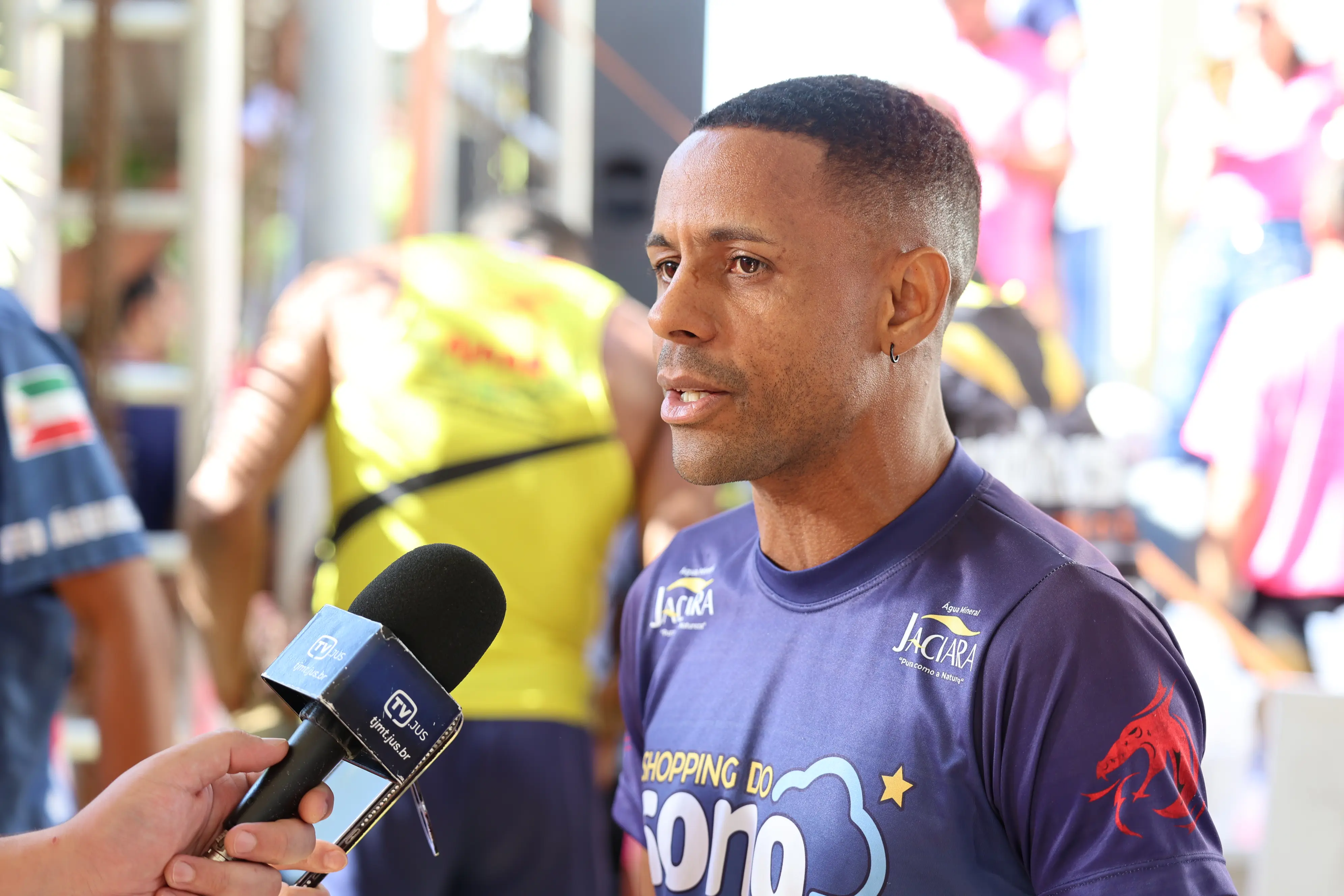 Um homem concede entrevista a microfone de emissora, durante evento esportivo. Veste camiseta estampada com patrocinadores da prova, em meio a outros participantes.