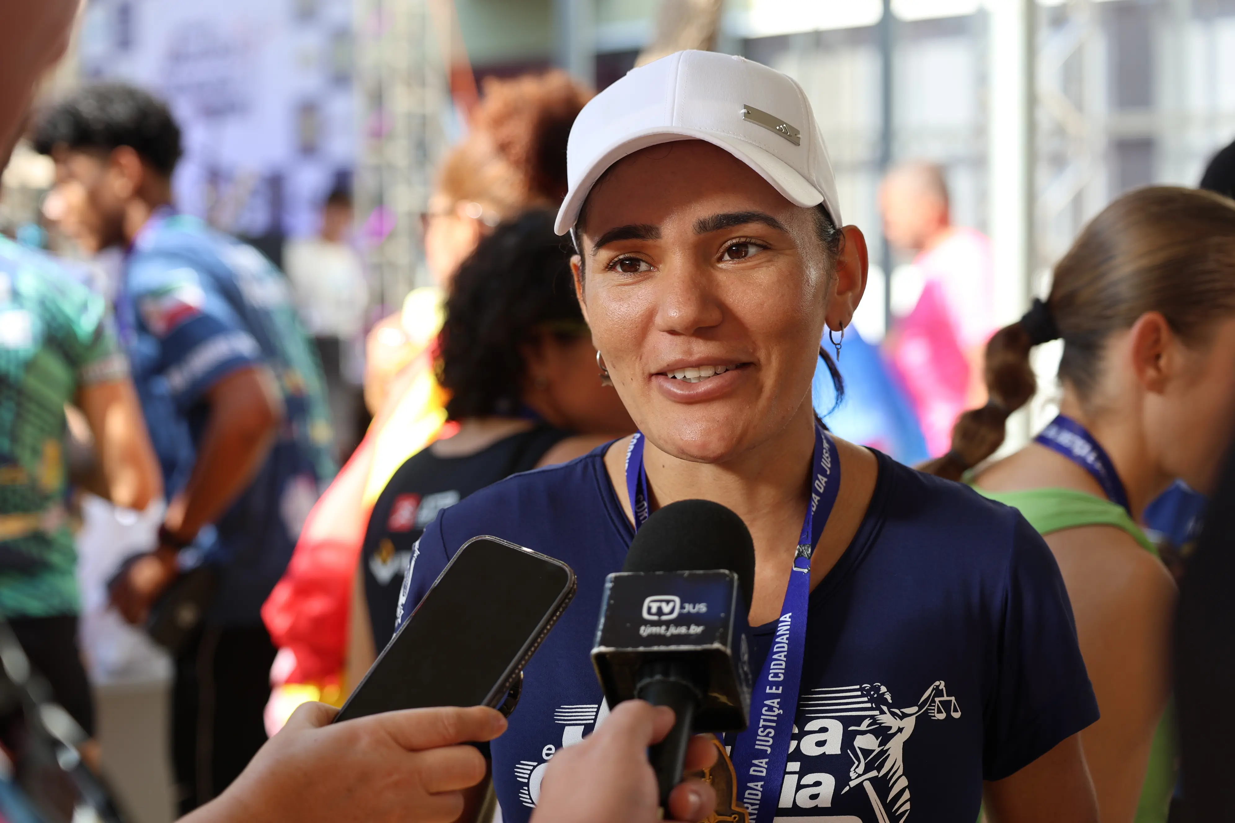 Uma mulher de boné branco sorri durante entrevista concedida a repórteres, que aproximam celular e microfone. Veste camiseta azul-marinho e exibe medalha ao pescoço.