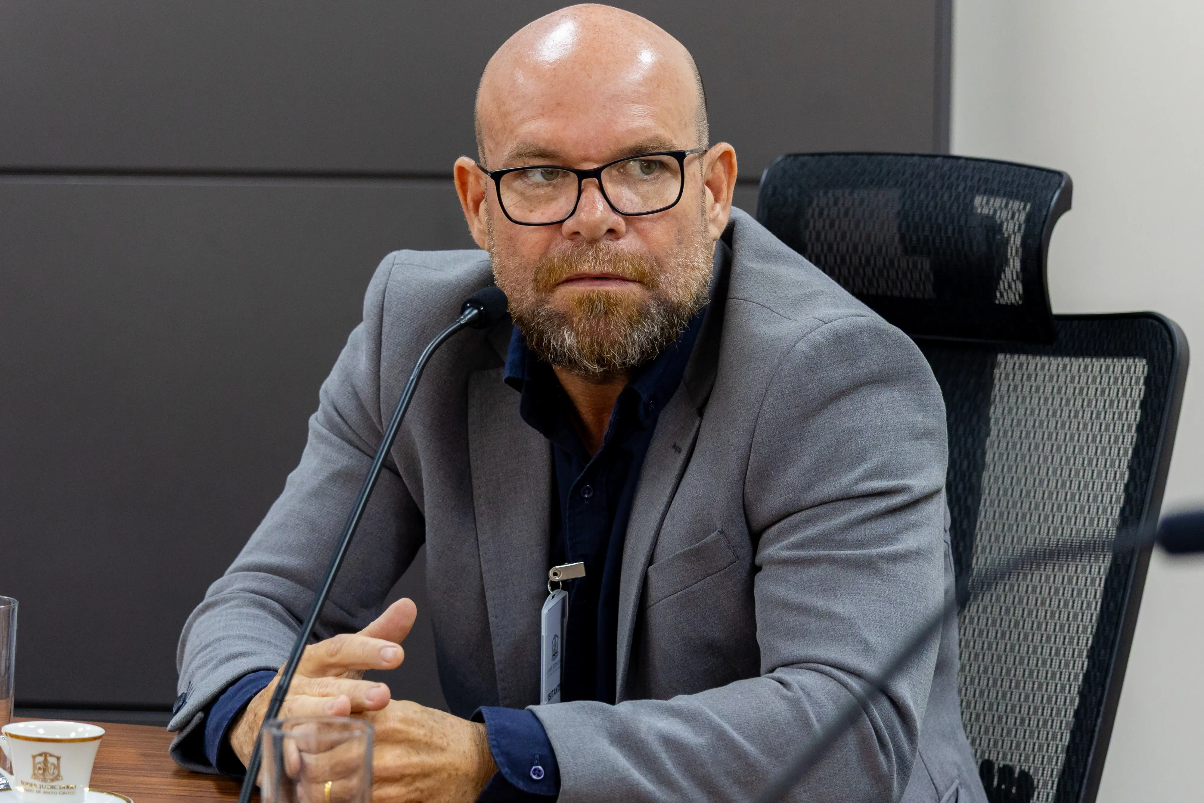 Homem careca, veste blazer cinza sobre camisa azul-marinho, usa óculos de armação preta e fala ao microfone. Xícara e copo d'água sobre a mesa.