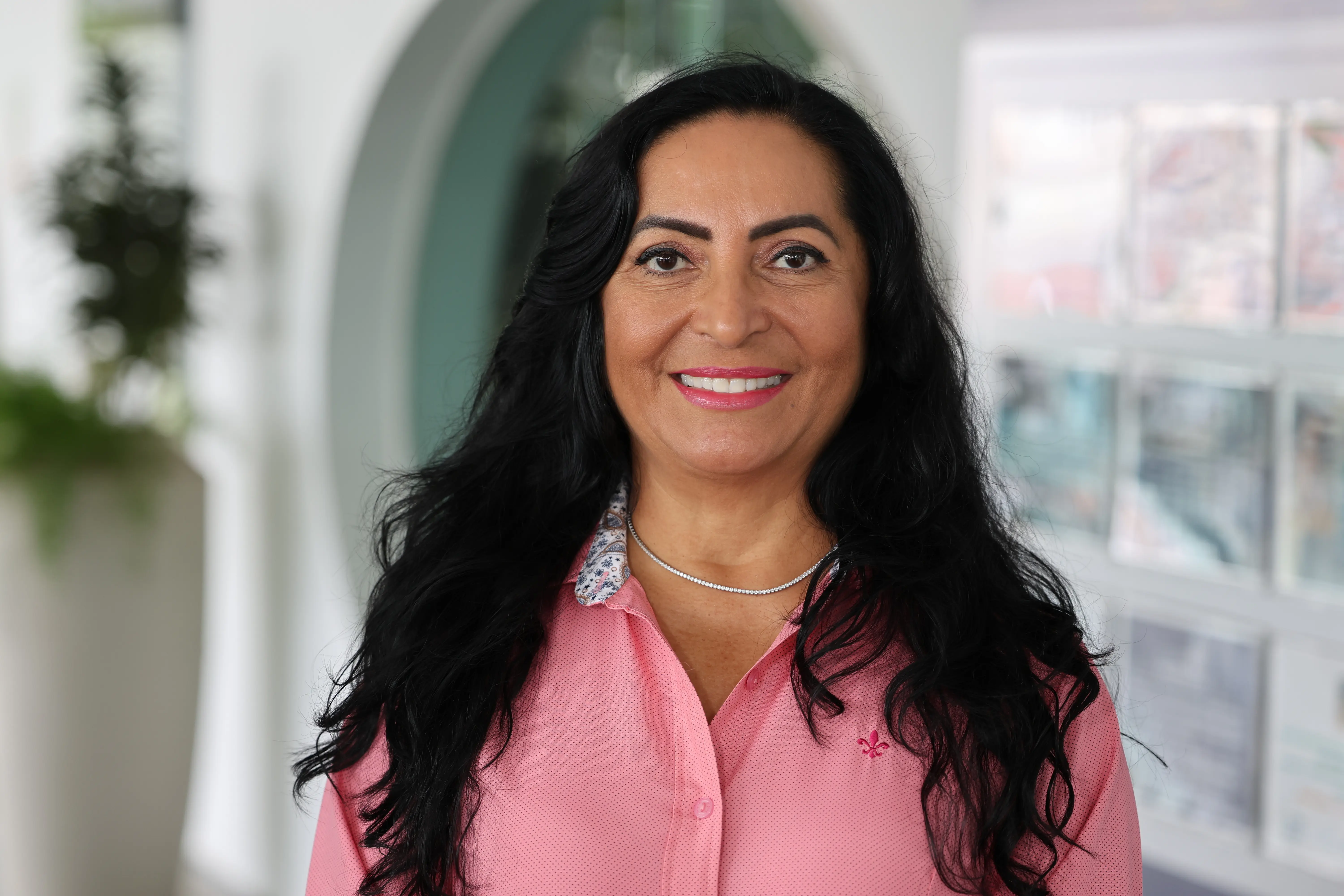 Retrato frontal da coordenadora Claudenice Deijany Farias de Costa. Ela está sorridente, tem longos cabelos pretos ondulados, veste uma camisa rosa e usa um colar fino prateado. O fundo está desfocado em tons claros.