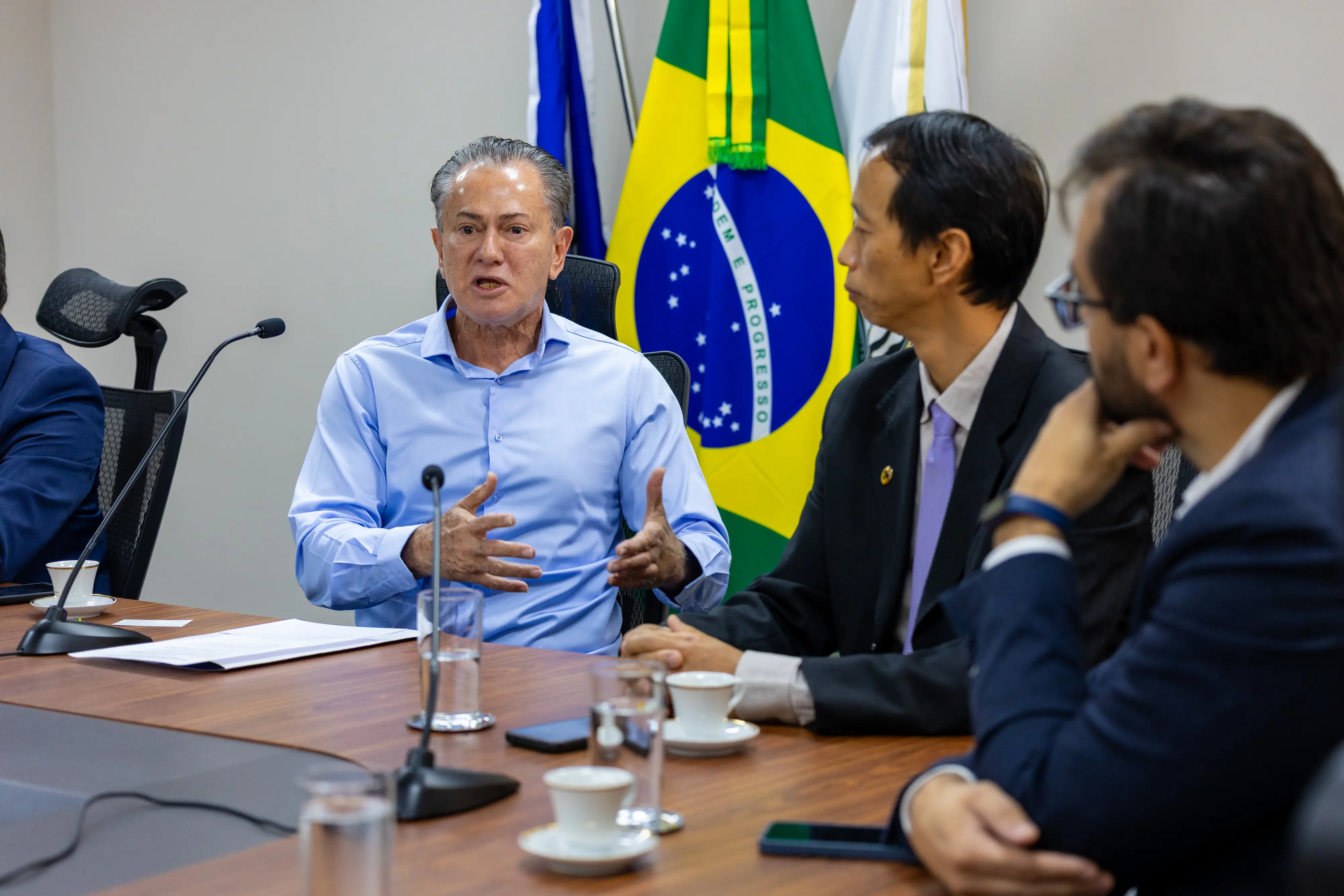 Homem de cabelos grisalhos, veste camisa social azul-claro, gesticula com as mãos enquanto fala. Ao fundo, as bandeiras do Brasil e do TJMT.