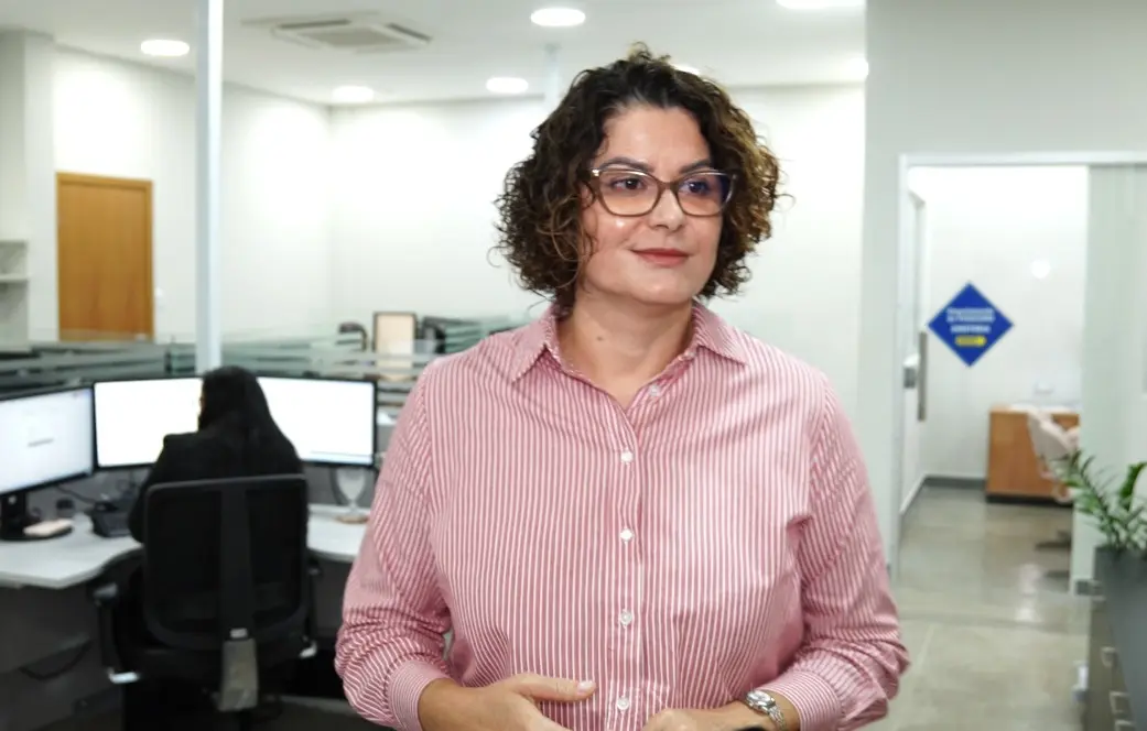 Foto horizontal colorida, em plano fechado, que mostra a gestora de Contabilidade do TJMT, Luzia Monteiro, em pé, sorrindo, na sala onde ela trabalha. Ela é uma mulher branca, de cabelos curtos, cacheados e castanhos, usando camisa social rosa clara listrada e óculos de grau.