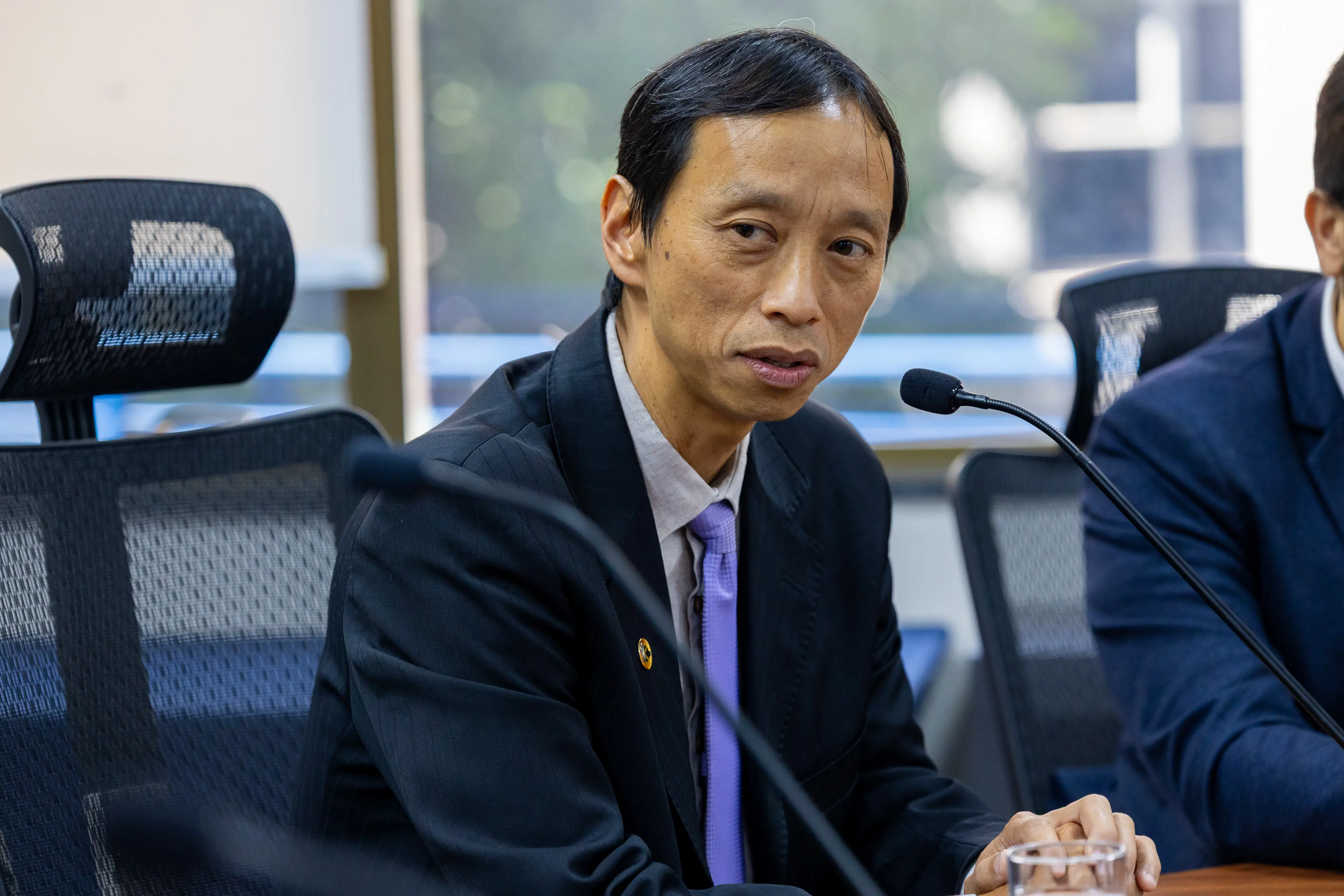 Homem oriental, de cabelos lisos escuros, veste terno preto e gravata lilás, fala ao microfone de mesa. Ao fundo, janelas com vegetação desfocada.
