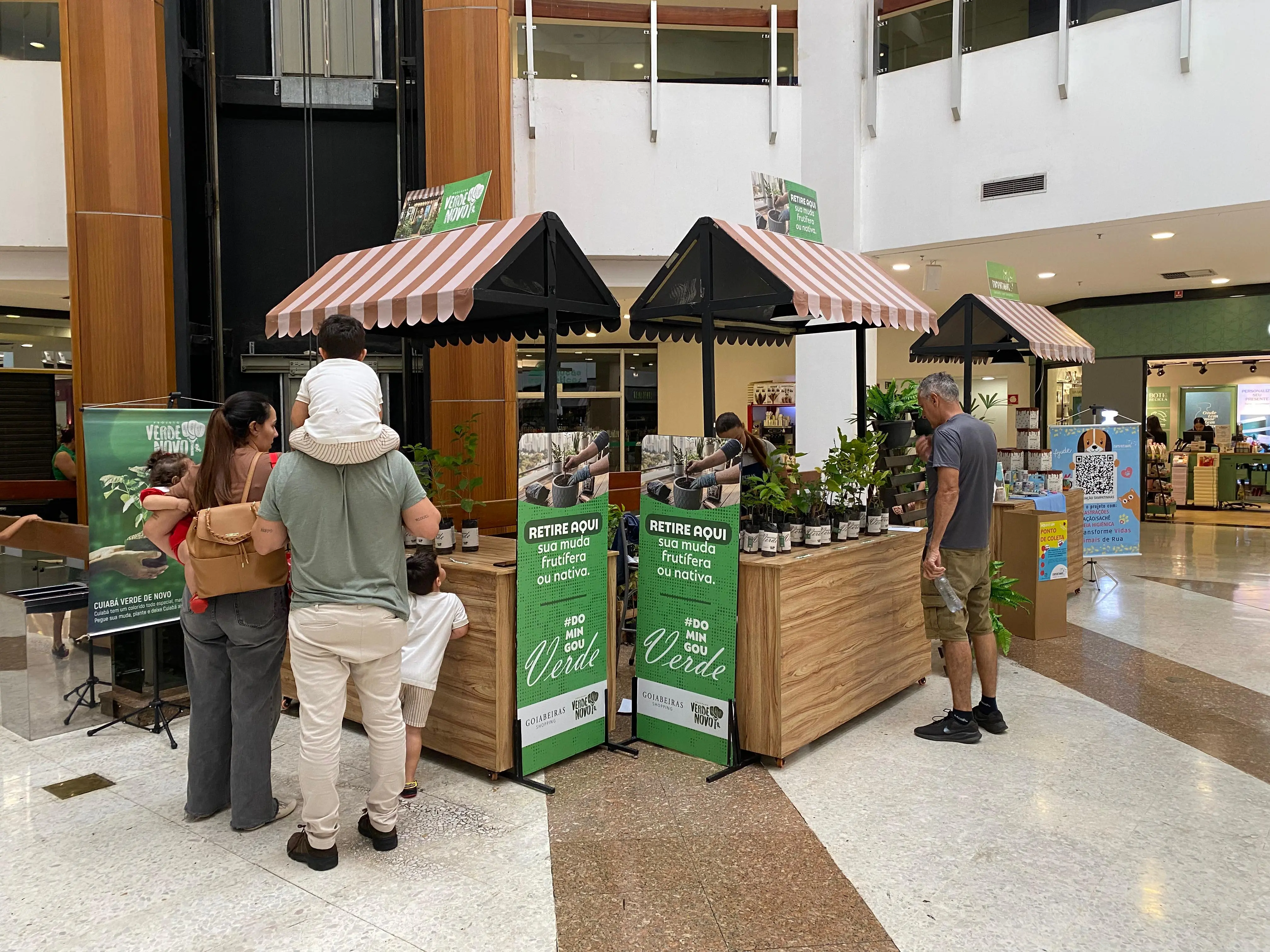 Estande de mudas de plantas em shopping. Barracas de madeira com coberturas listradas distribuem vasos. Família e homem olham. Placas verdes grandes anunciam evento "#DOMINGOU VERDE".