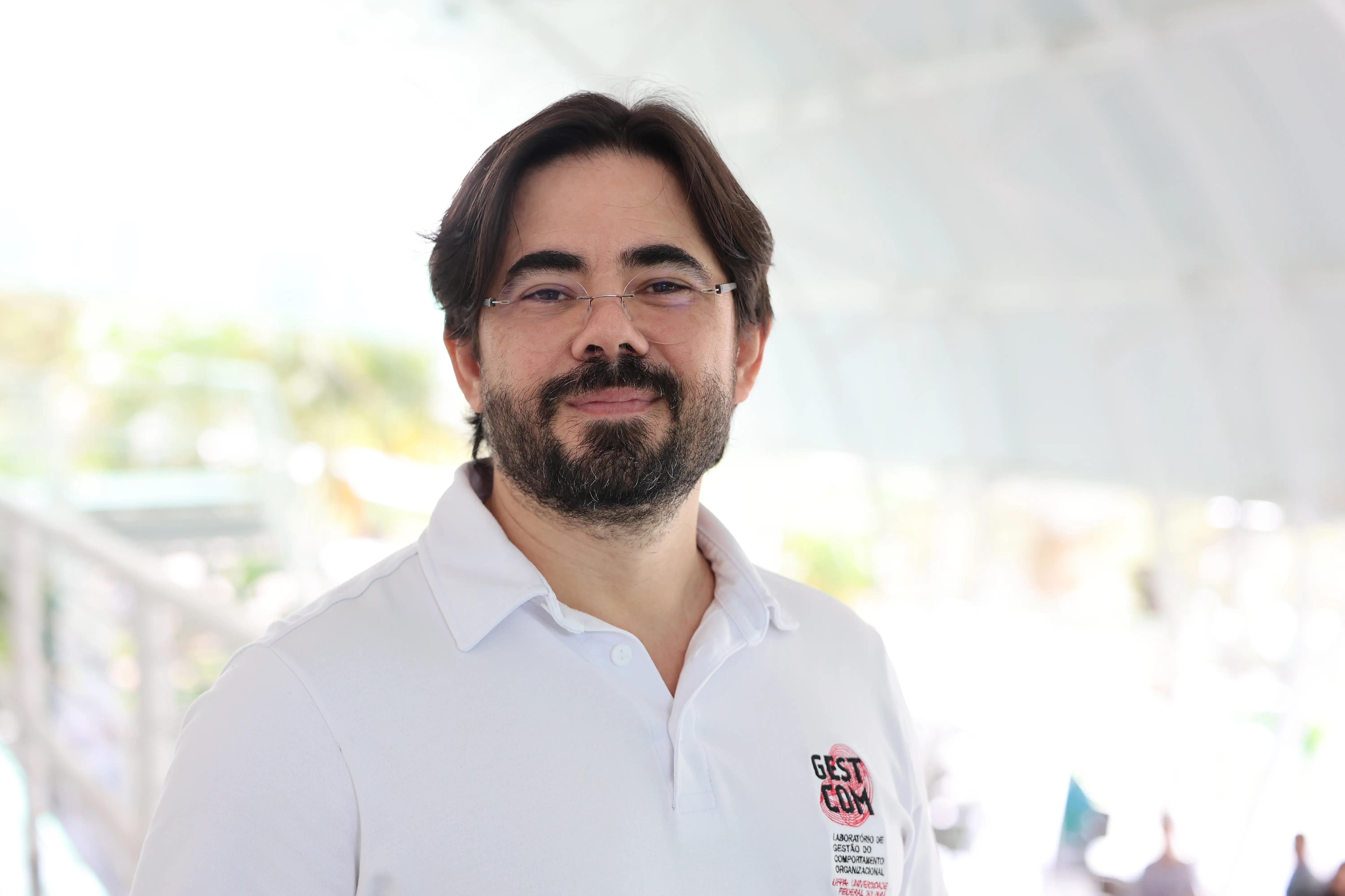Fotografia do professor Thiago Dias Costa. Ele usa óculos, tem barba e cabelos castanhos, veste uma camisa polo branca com um pequeno logotipo no lado esquerdo do peito. 