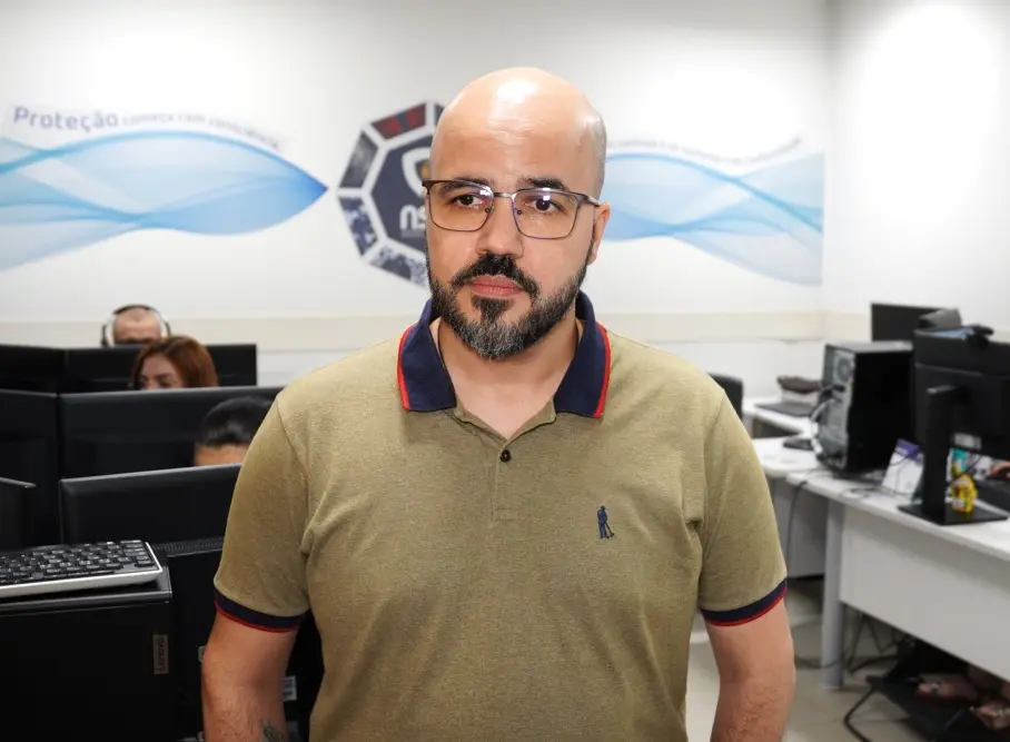 Foto horizontal colorida, em plano fechado, que mostra Rafael Nogueira, gestor do Núcleo de Inteligência Artificial do TJMT, em pé, na sala do núcleo. Ele é um homem jovem, branco, careca, de olhos e barba escuros, usando camiseta polo verde com gola azul e óculos de grau.