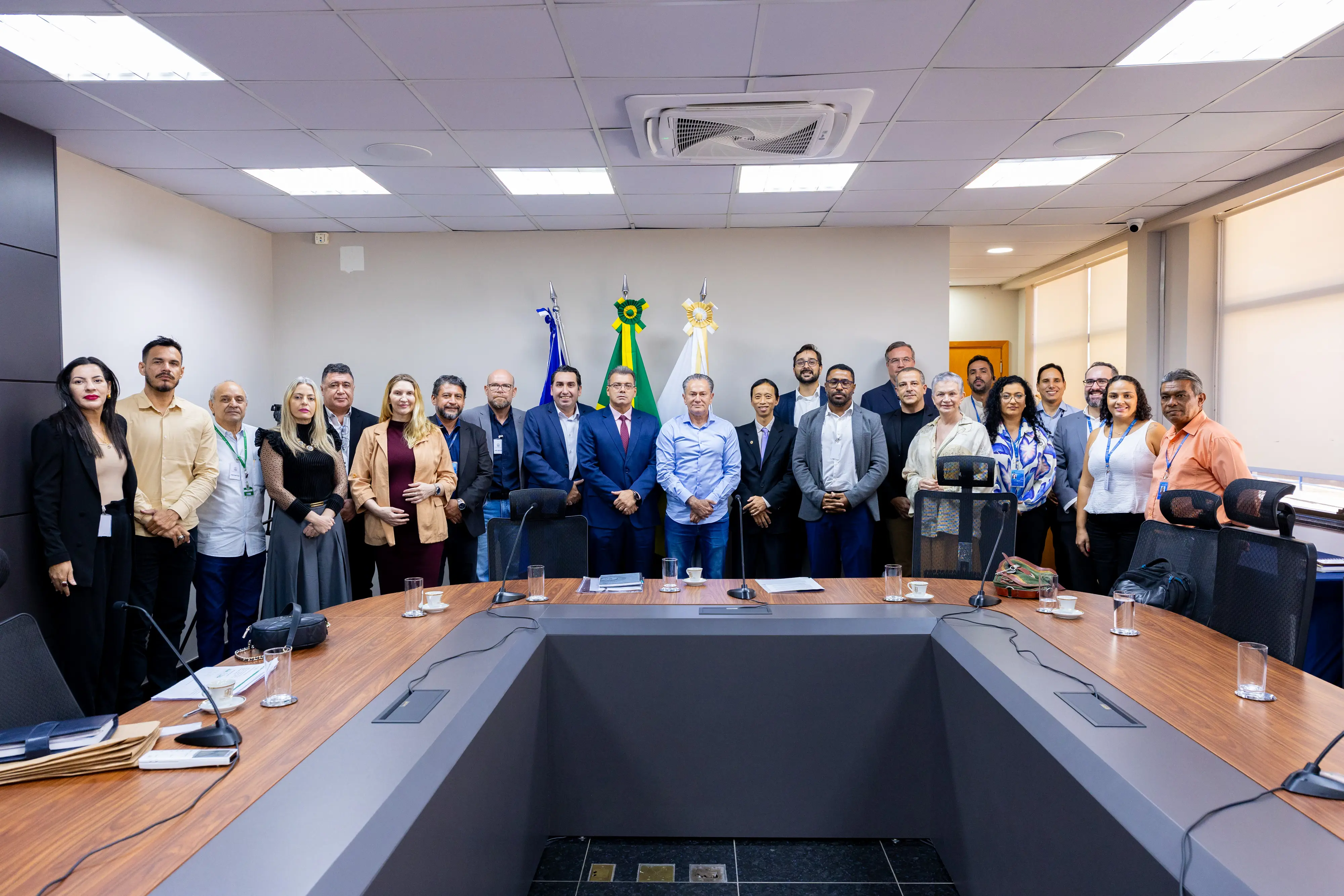 Grupo de aproximadamente 25 pessoas posadas em semicírculo atrás de mesa oval em sala institucional. Ao fundo, bandeiras do Brasil e do TJMT.