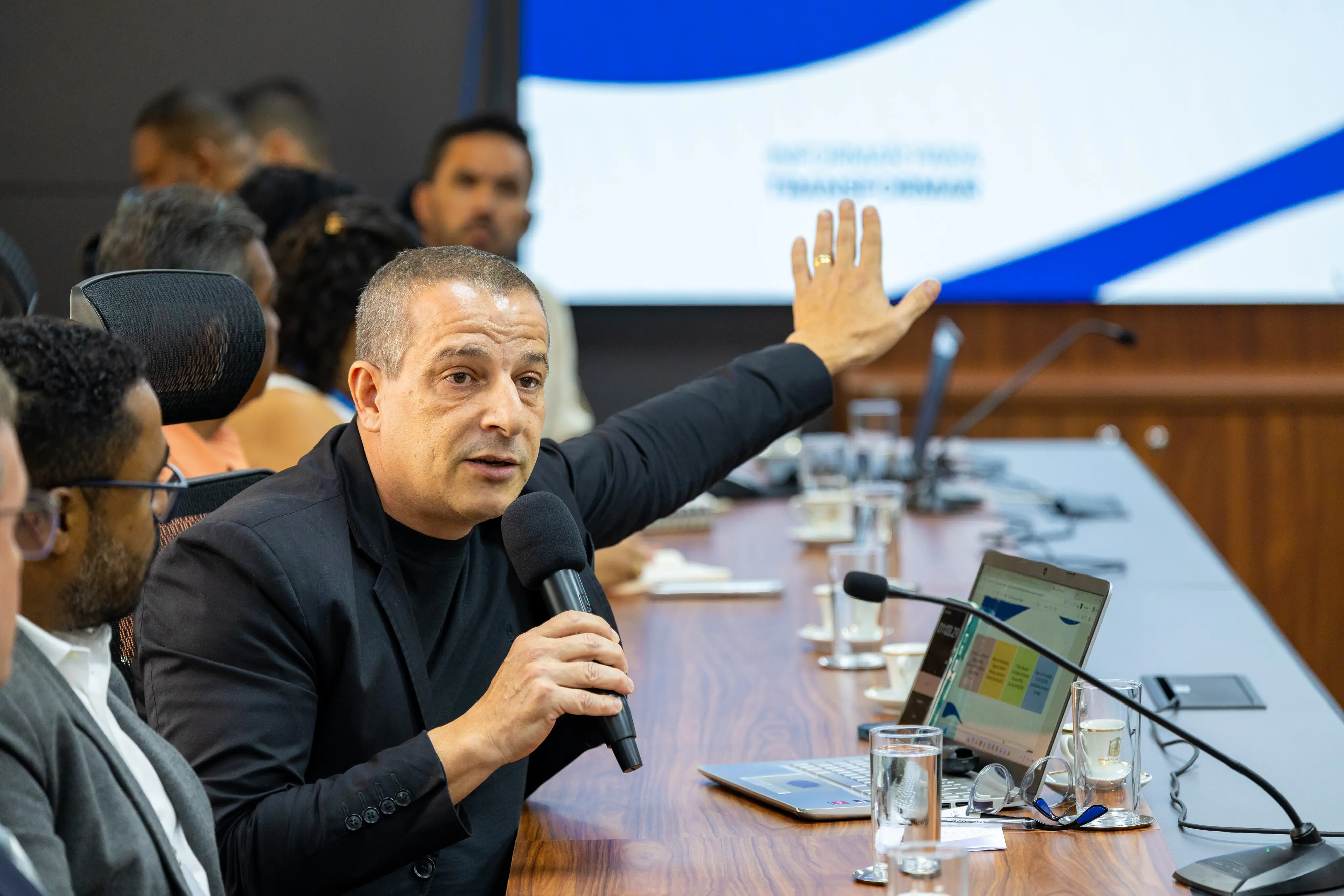 Homem careca, veste conjunto preto, segura microfone e levanta o braço. Ao fundo, tela azul e branca exibe uma apresentação. Mesa longa com laptops e copos d'água.