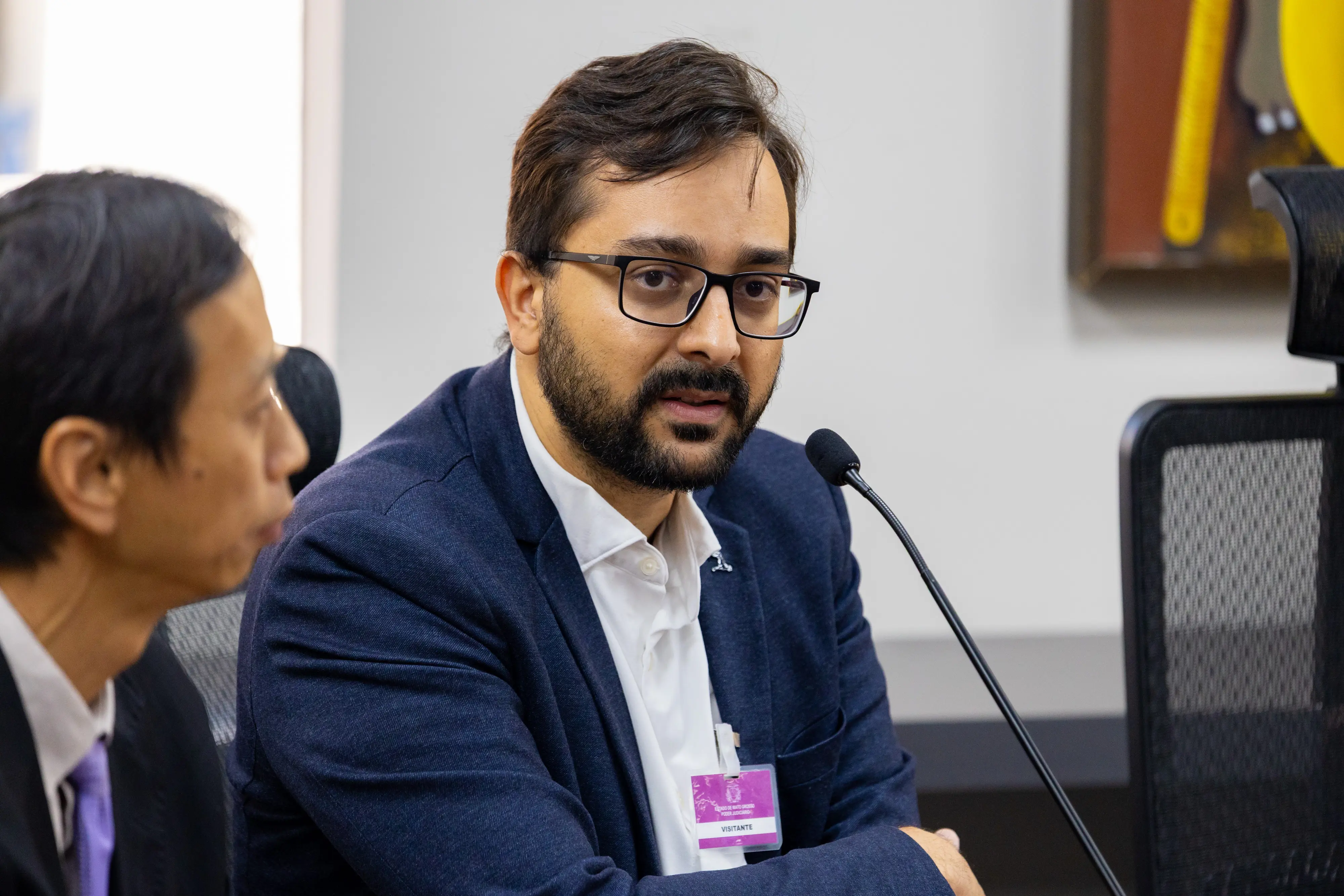 Homem de cabelos escuros e barba curta, veste blazer azul-marinho sobre camisa branca, fala ao microfone. Ao fundo, outro homem aparece desfocado à esquerda.