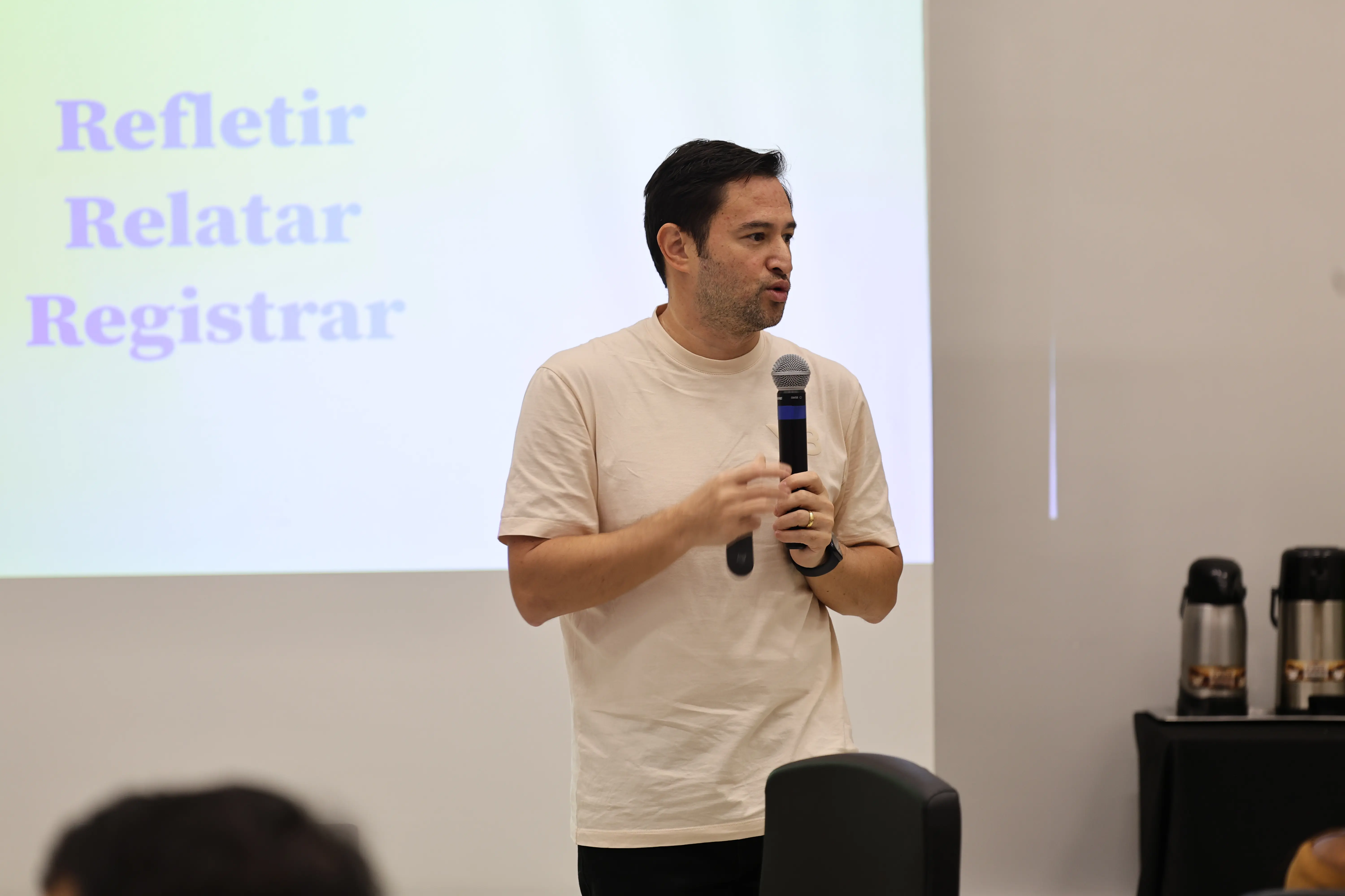 Homem jovem de camiseta clara fala ao microfone diante de uma tela de projeção com as palavras "Refletir, Relatar, Registrar". Ao fundo, garrafas térmicas sobre uma mesa lateral.