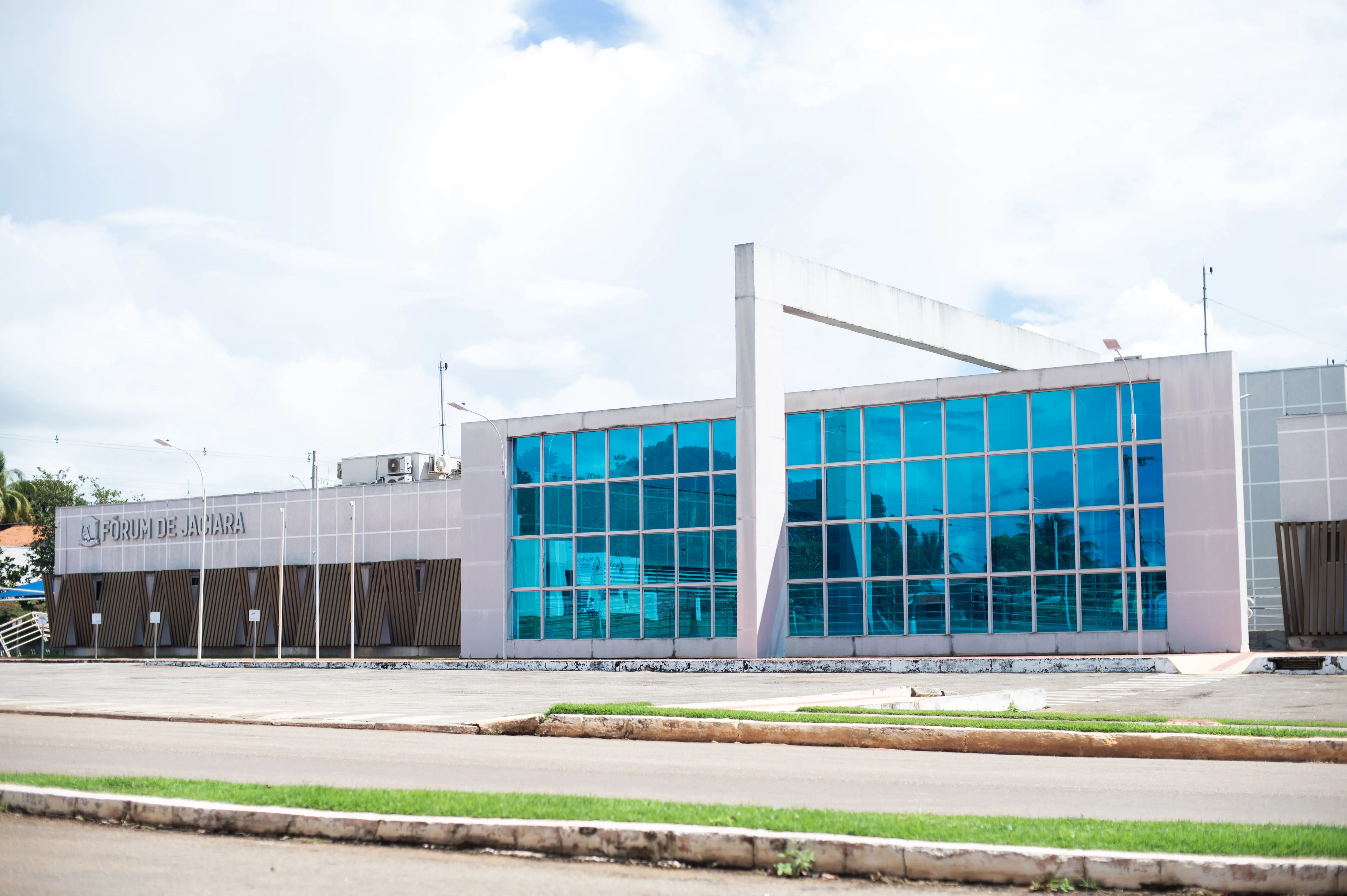 Imagem mostra a fachada moderna do Fórum de Jaciara, com paredes de vidro azul espelhado, estrutura clara e ampla área externa asfaltada, sob um céu parcialmente nublado.