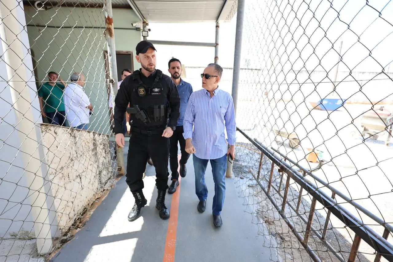 Homem de camisa listrada azul e branca, acompanhado de policial penal e outro homem, caminha por corredor externo cercado por tela metálica, em área de unidade prisional sob sol forte.