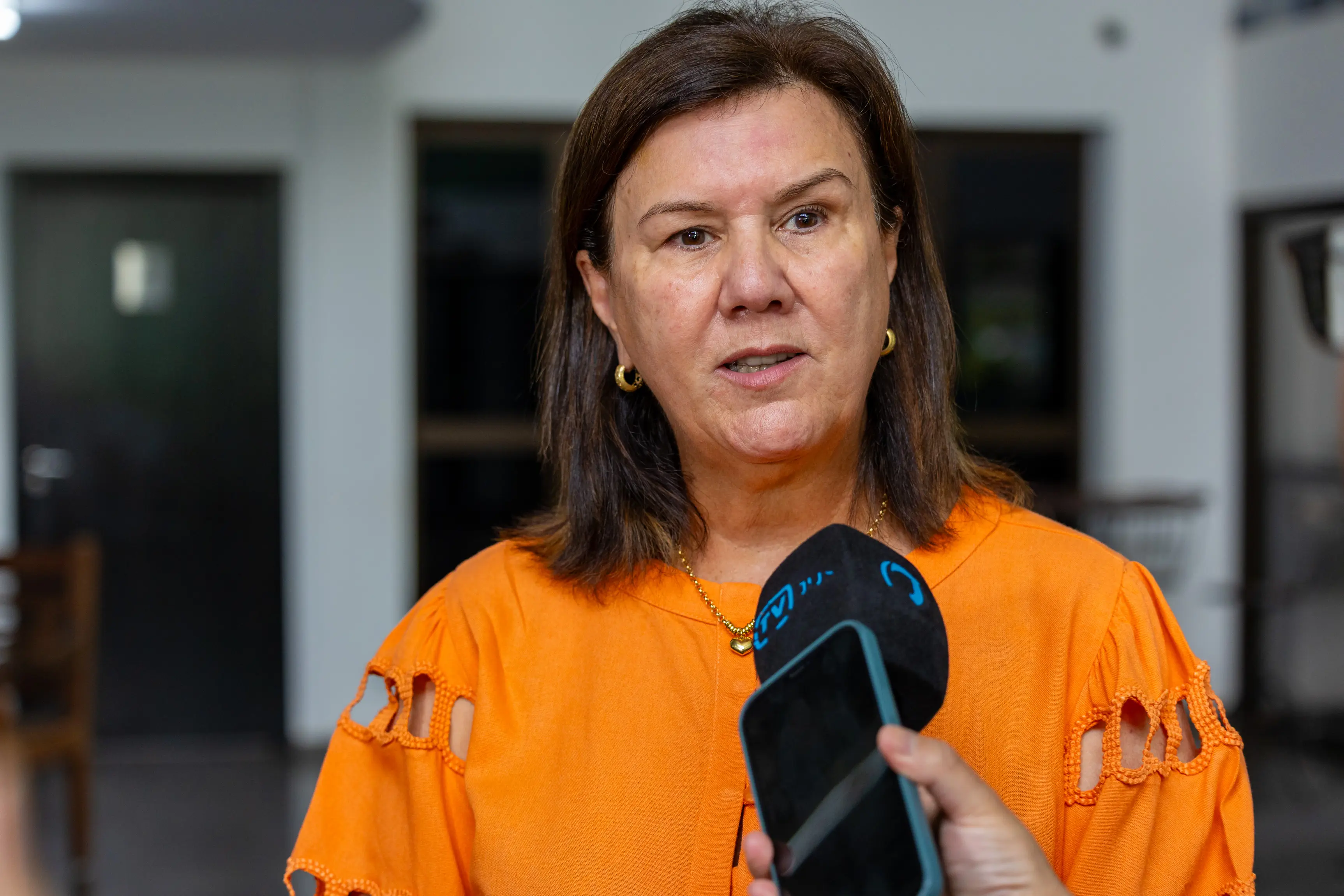 Mulher de pele clara e cabelos castanhos, vestindo roupa laranja com detalhes vazados nas mangas, fala para um microfone preto e um celular. O fundo da imagem está desfocado em tons de cinza.