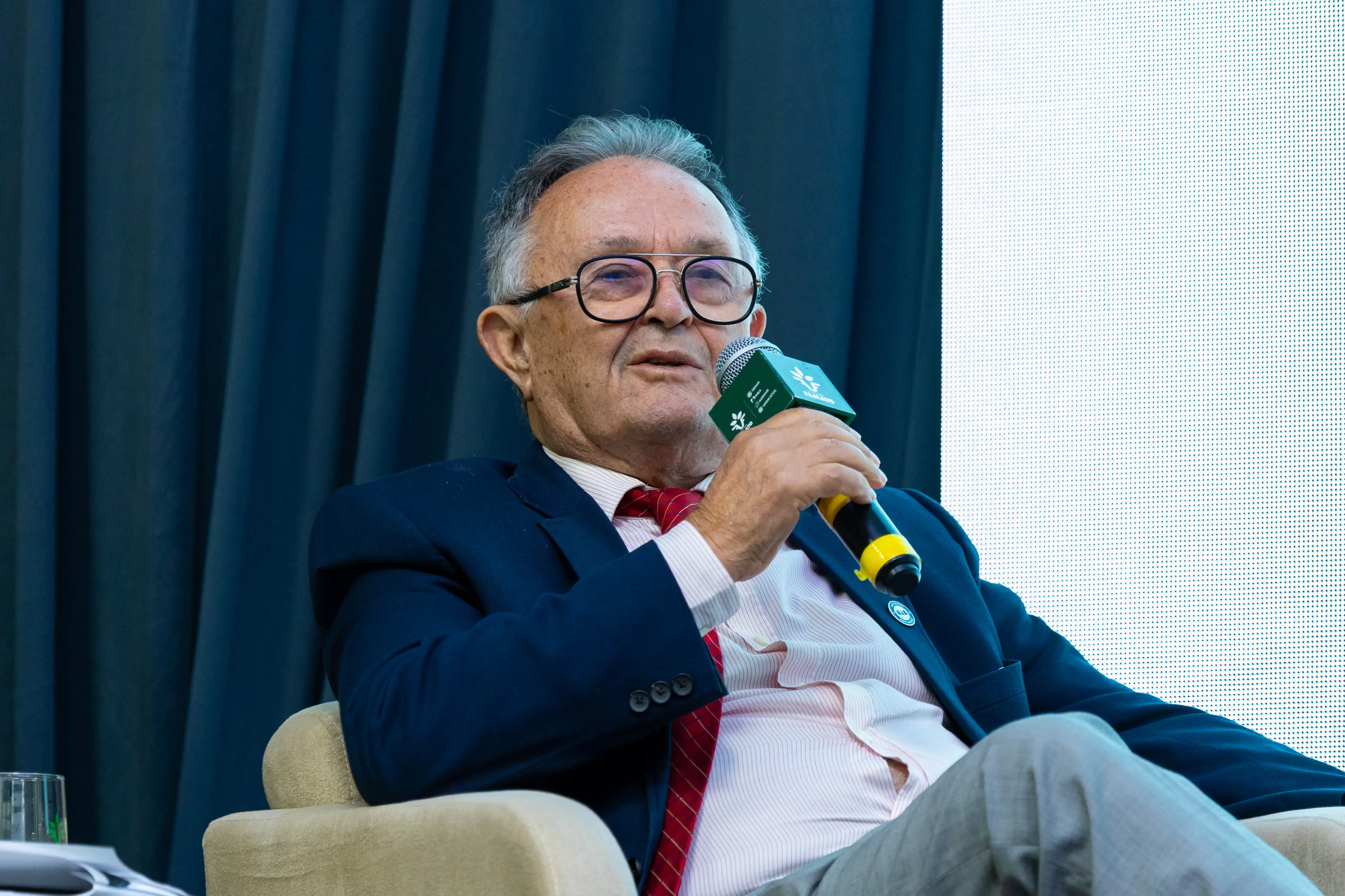 Um homem idoso de óculos e terno azul fala ao microfone em um evento. Ele está sentado em uma poltrona clara, com uma cortina azul ao fundo. O microfone possui uma espuma verde com logotipos.
