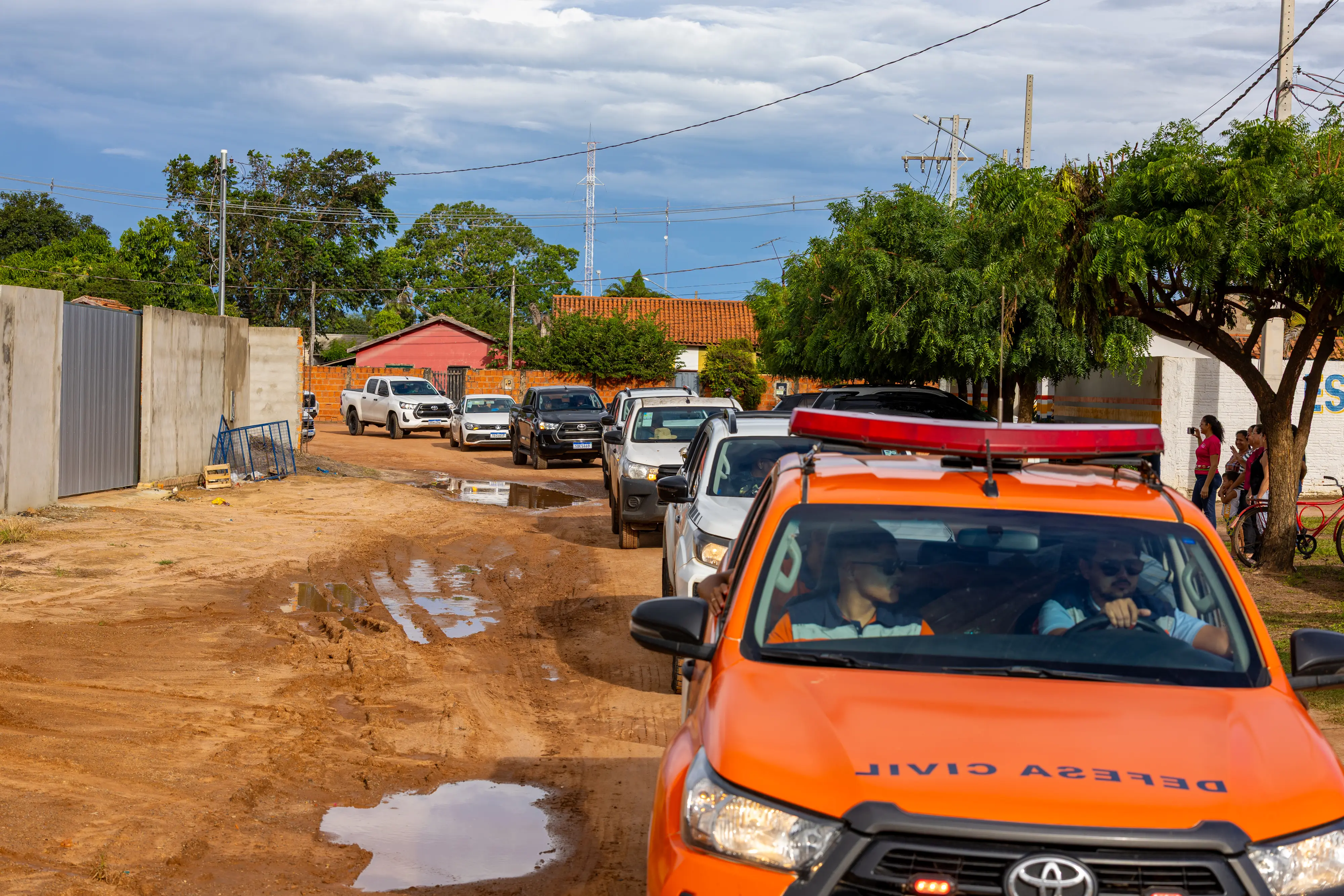 Comboio de veículos em estrada de terra com poças. Em destaque, caminhonete laranja da Defesa Civil lidera a fila, seguida por carros brancos e pretos sob céu nublado.
