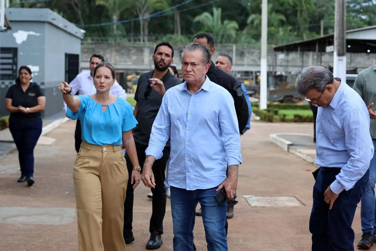 Uma mulher de blusa azul aponta para frente enquanto caminha ao lado de um homem de camisa social azul clara e calça jeans. Eles estão em uma área externa pavimentada com outras pessoas ao fundo.