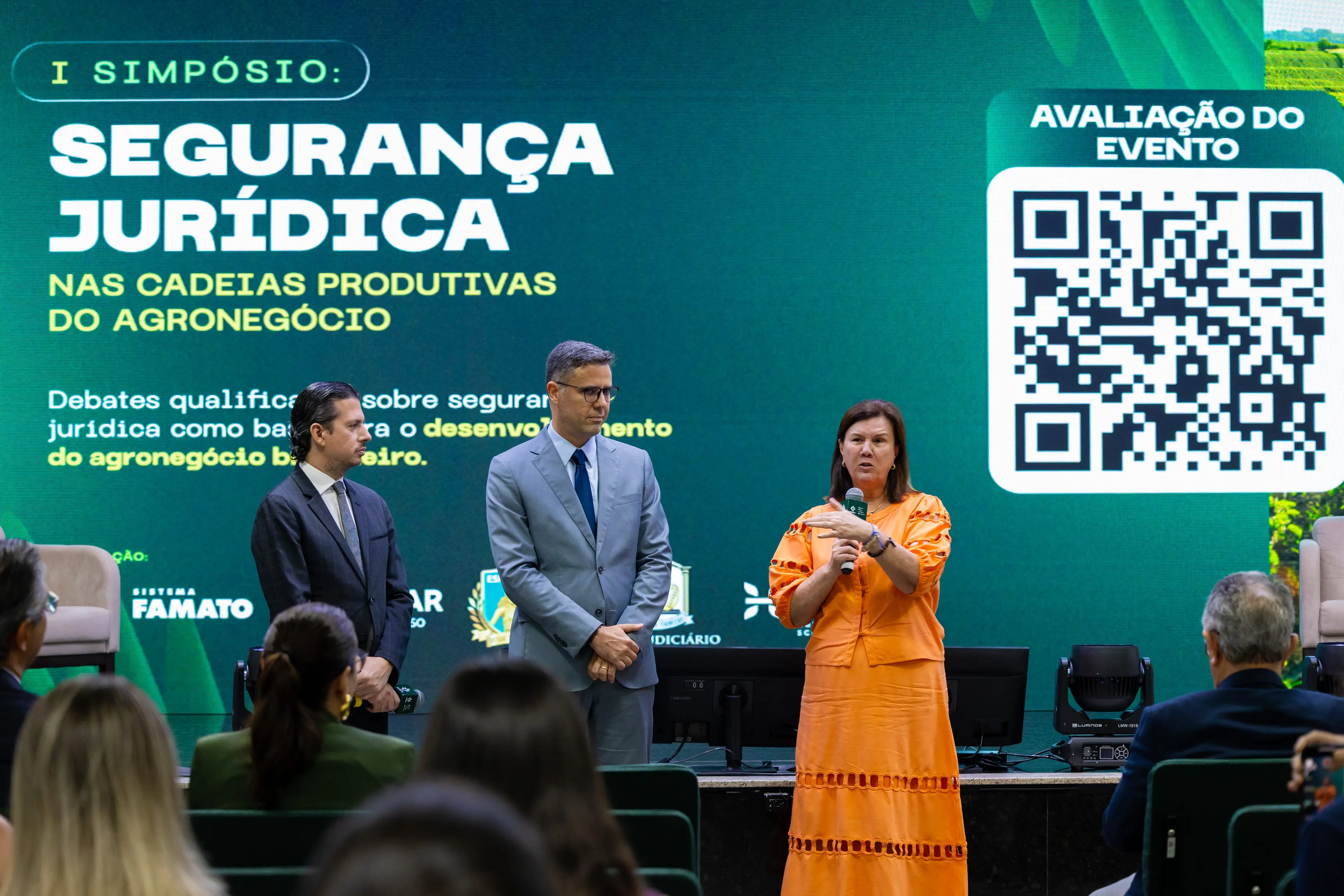 Uma mulher de vestido laranja discursa. Ao lado, dois homens de terno observam. Ao fundo, um telão verde exibe 