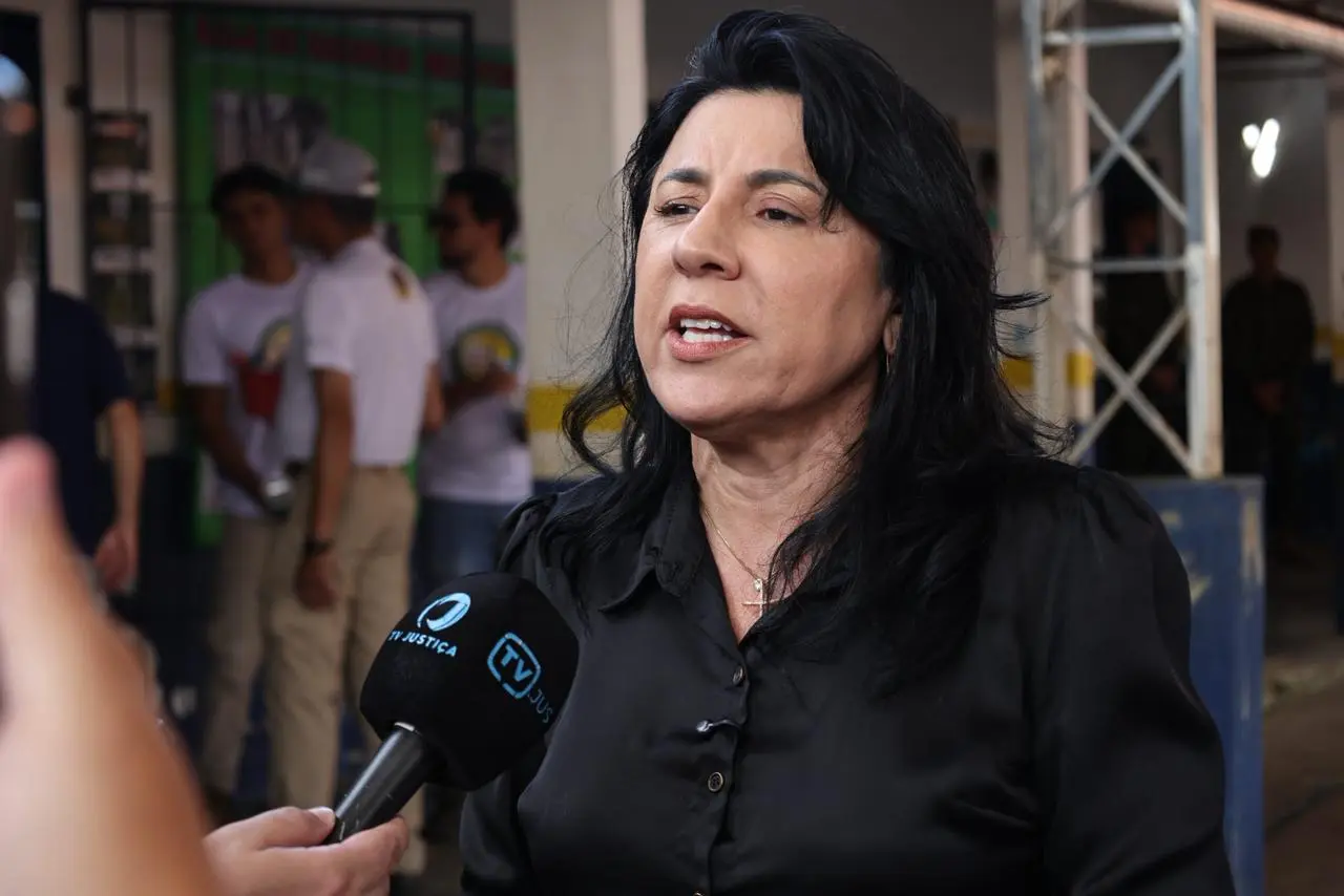 Mulher de cabelos escuros e blusa preta fala ao microfone da TV Justiça. Ela tem expressão séria e decidida. Ao fundo, de forma desfocada, pessoas e uma estrutura metálica em ambiente externo.