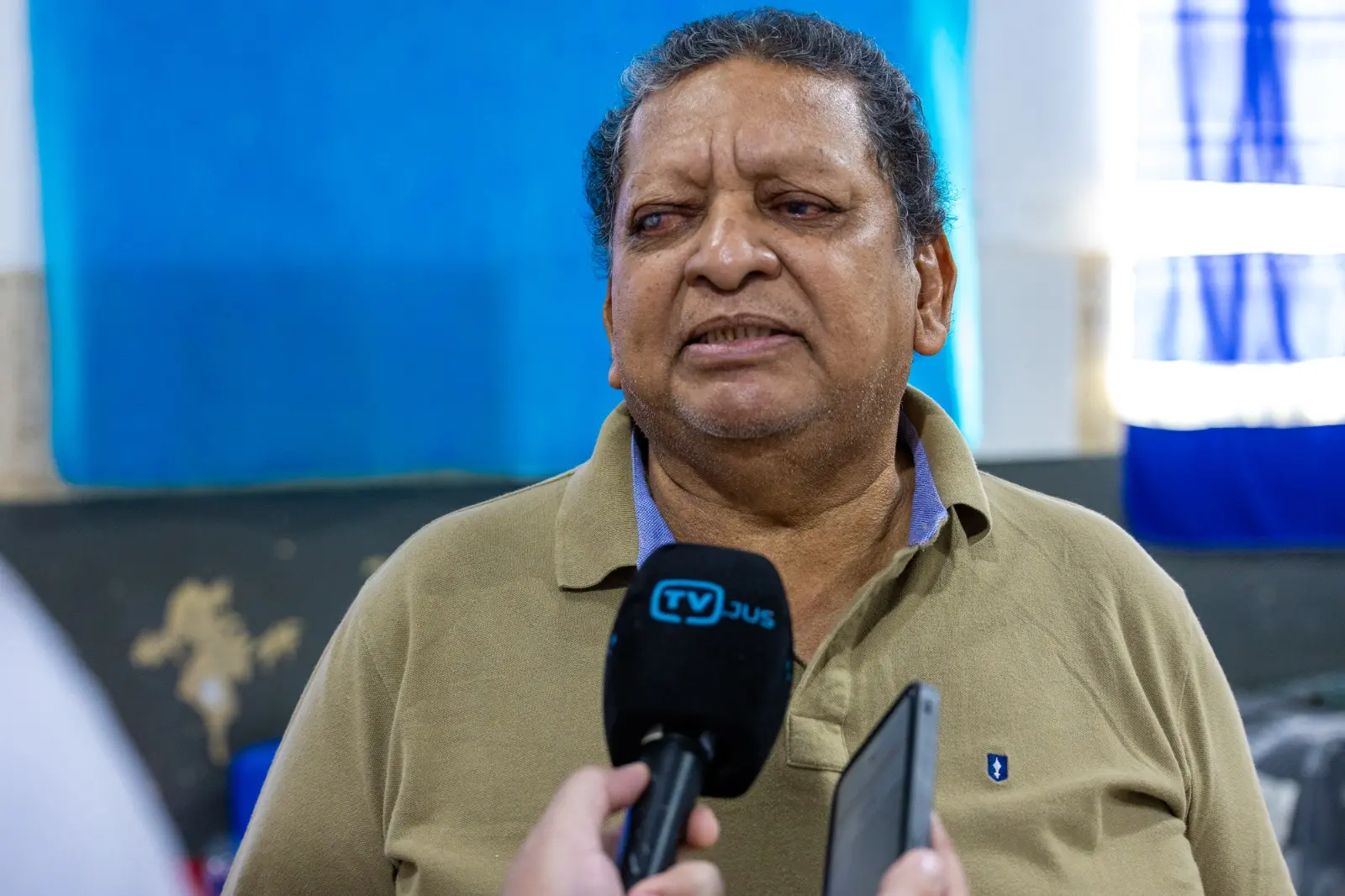 Homem de meia-idade concede entrevista à TV Justiça. Ele usa camisa polo bege e fala ao microfone enquanto segura um celular, relatando informações ou avaliando os atendimentos prestados na ação social.