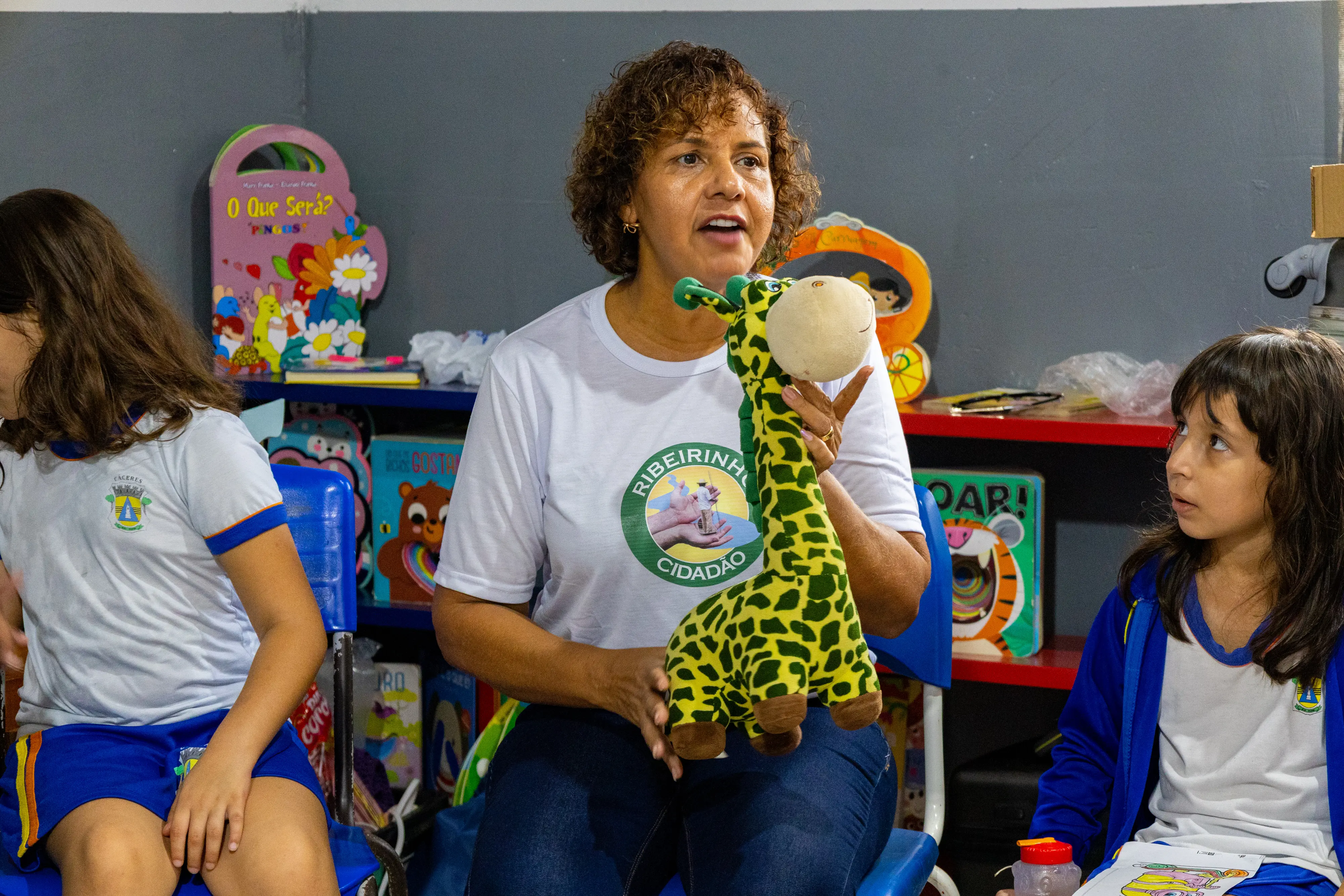 Mulher de cabelos cacheados e camiseta branca do projeto Ribeirinho Cidadão segura uma girafa de pelúcia entre duas crianças sentadas. Ao fundo, uma estante com livros e materiais lúdicos.