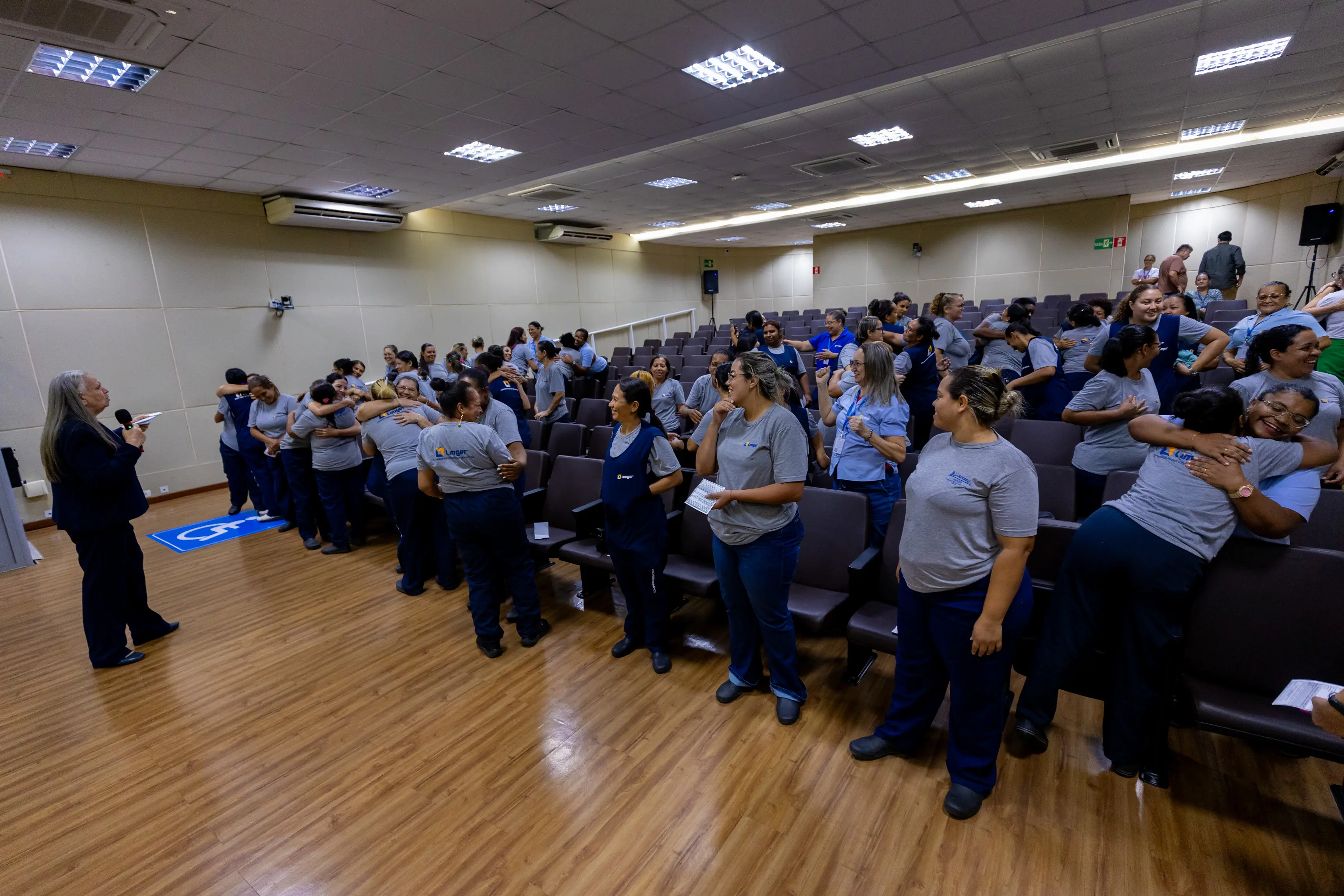 Auditório cheio com dezenas de pessoas sentadas, a maioria usando camisetas azuis. Participantes levantam as mãos e interagem com quem conduz a atividade na frente da sala, em momento de dinâmica coletiva.