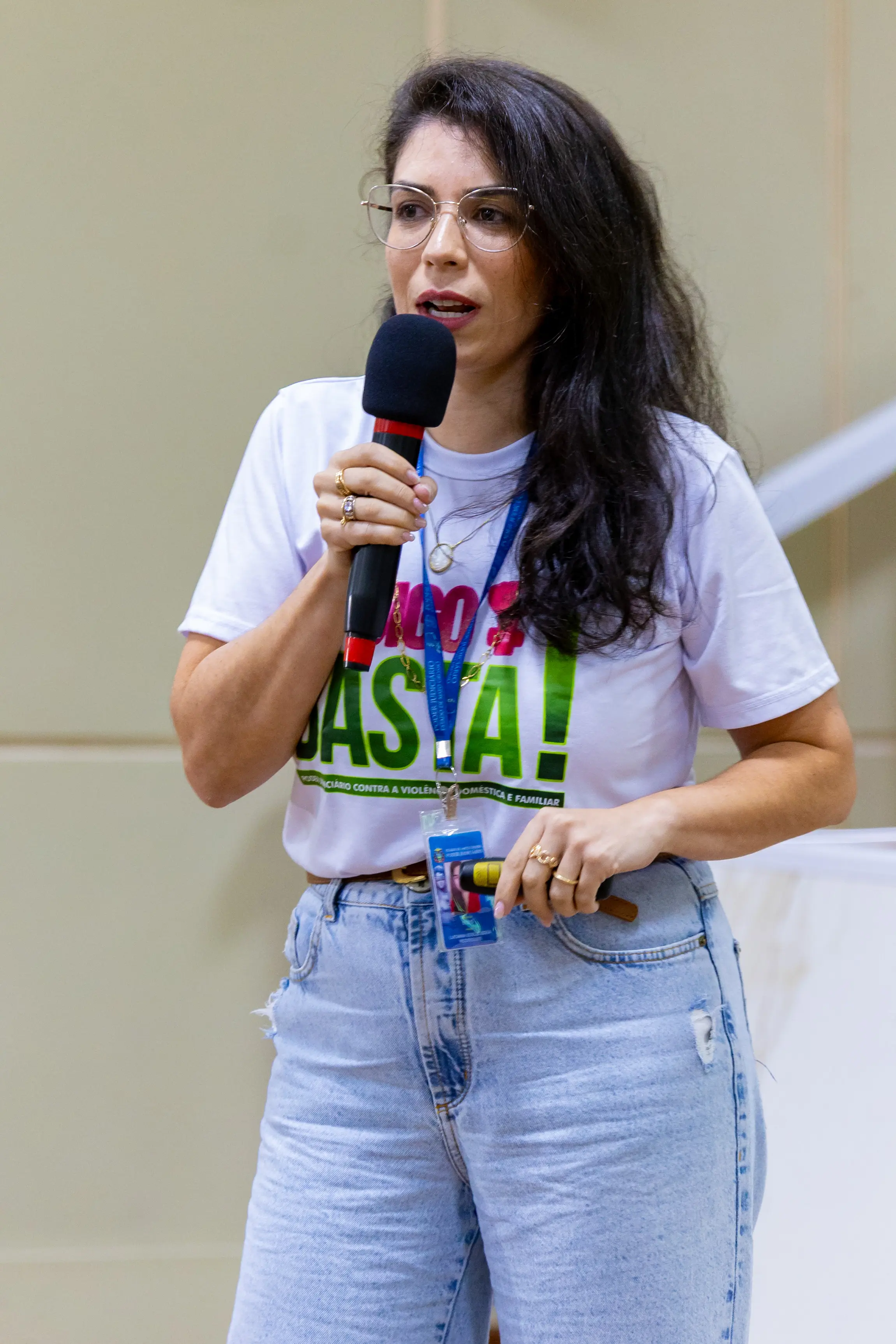 Close da parte superior de uma mulher segurando um microfone durante fala em evento. Ela veste camiseta branca com estampa colorida escrita “Viva a Justiça!”. O foco está no microfone e na mensagem da camiseta.