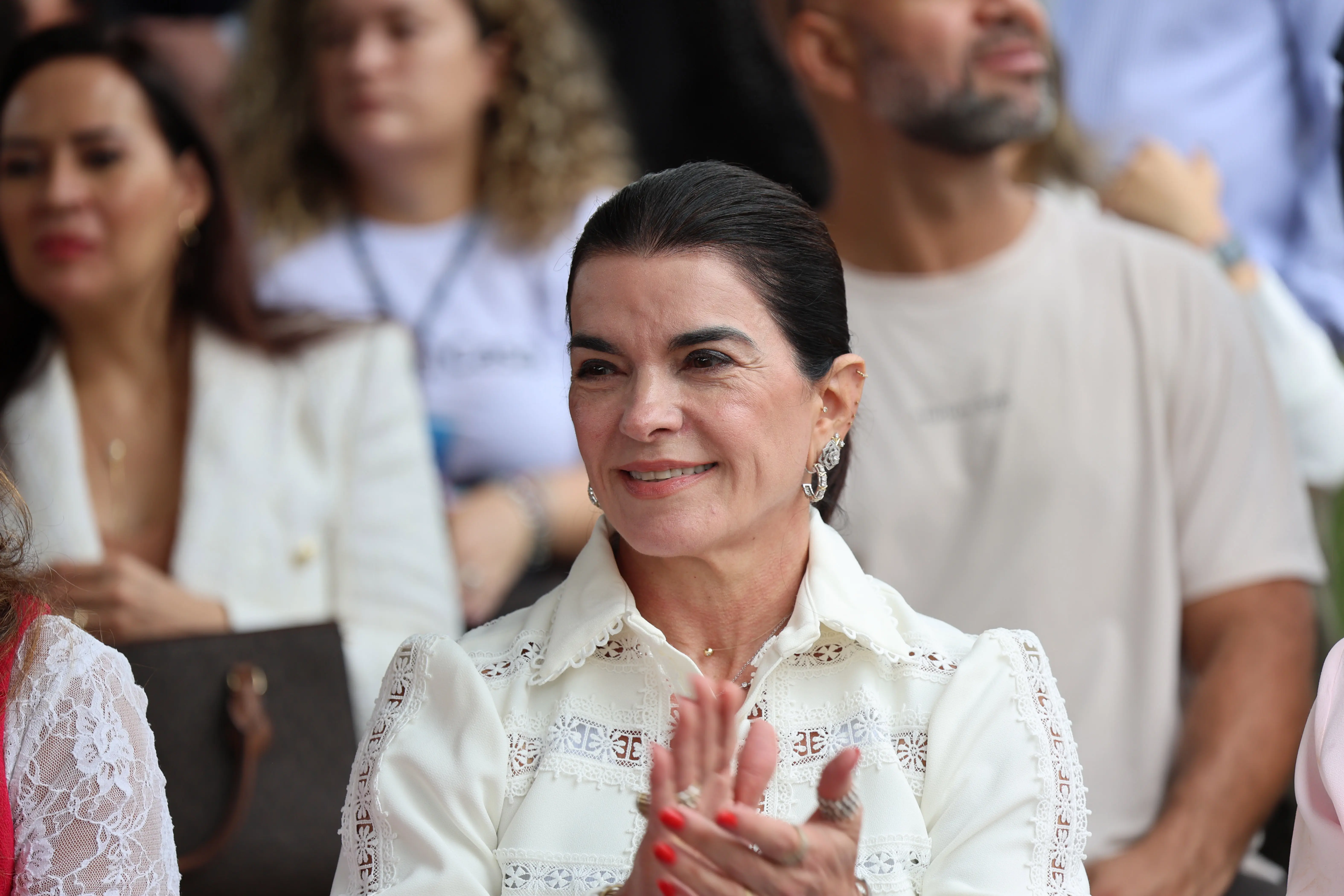 Juíza Eulice Jaqueline Cherulli, mulher branca de cabelos pretos presos. Sorri levemente e aplaude. Veste blusa branca com detalhes em renda e gola trabalhada. Usa brincos de argola com brilho.