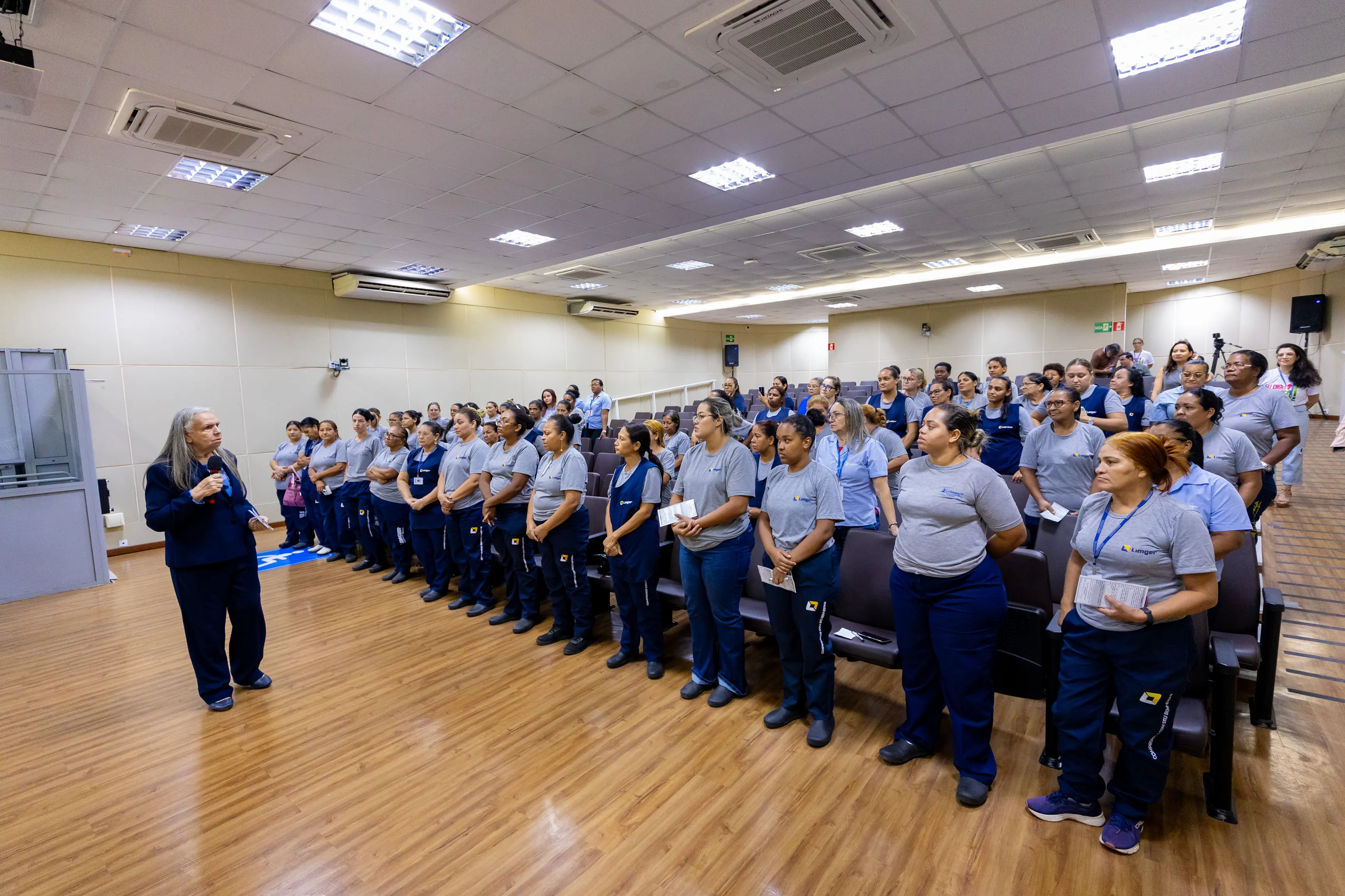 Vista ampla do auditório mostra dezenas de participantes em pé, organizados em fileiras. Uma facilitadora conduz a atividade à frente da sala enquanto o grupo acompanha atento durante momento coletivo.