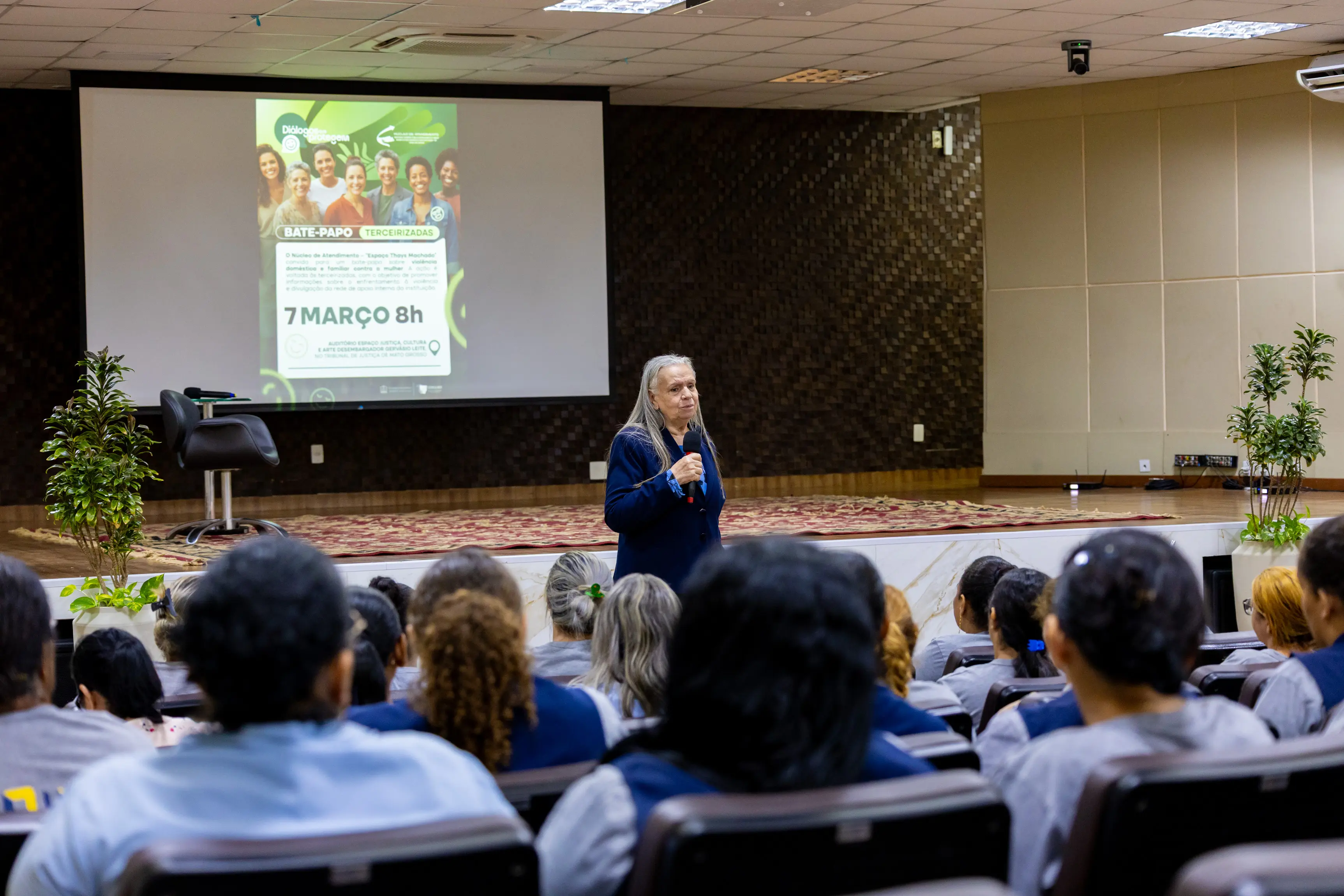 Desembargadora Maria Erotides conduz apresentação diante de um grande auditório cheio. O público está sentado em fileiras acompanhando a palestra, enquanto o telão exibe conteúdo do encontro.