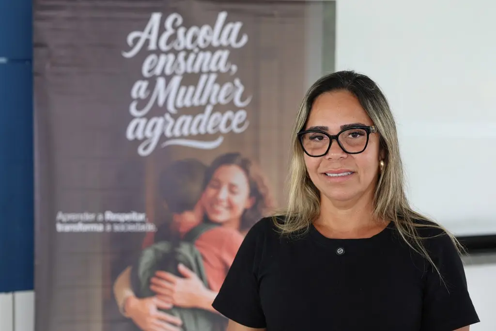 Foto horizontal em plano médio que mostra a diretora escolar, Elma Francisca Gomes, sorrindo para a foto, na frente de um banner da Cemulher, onde está escrito 