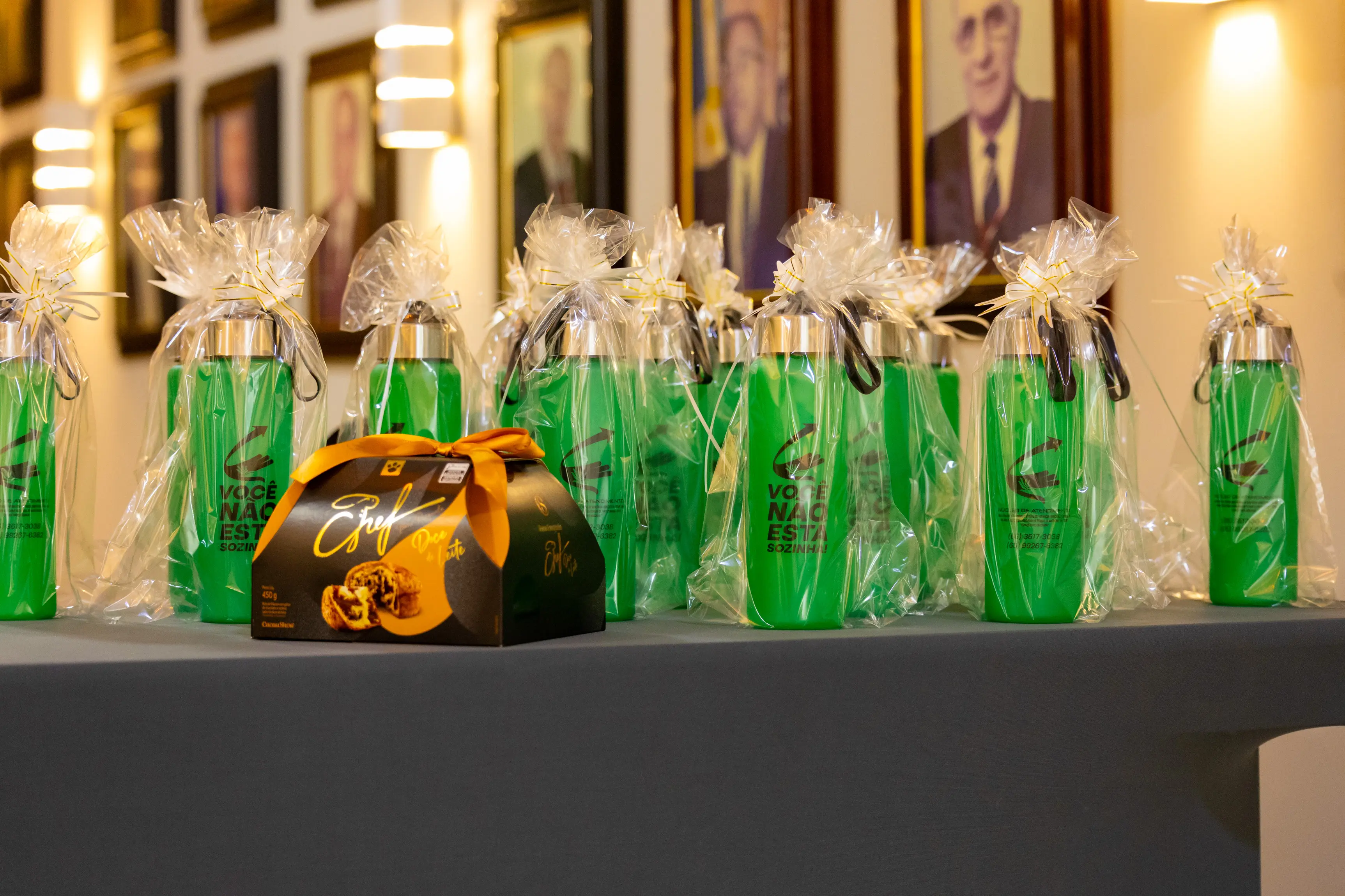 Mesa expositiva apresenta várias garrafas e frascos de vidro nas cores verde e âmbar, iluminados por luz suave. Os objetos decorativos compõem cenário utilizado durante atividade ou ambientação do evento.