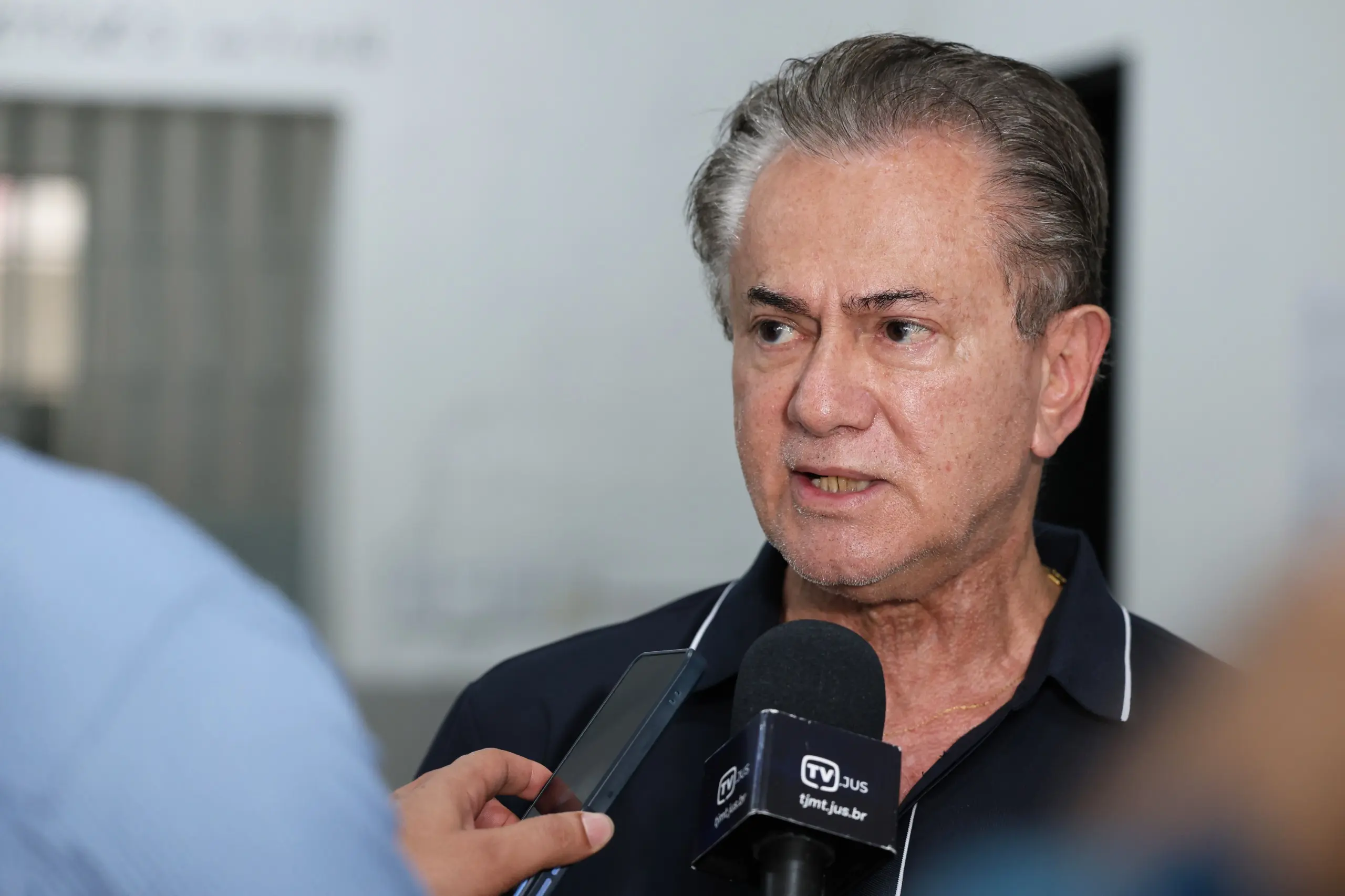 Retrato de um homem maduro de cabelos grisalhos e camisa polo azul marinho. Ele fala para um microfone da 