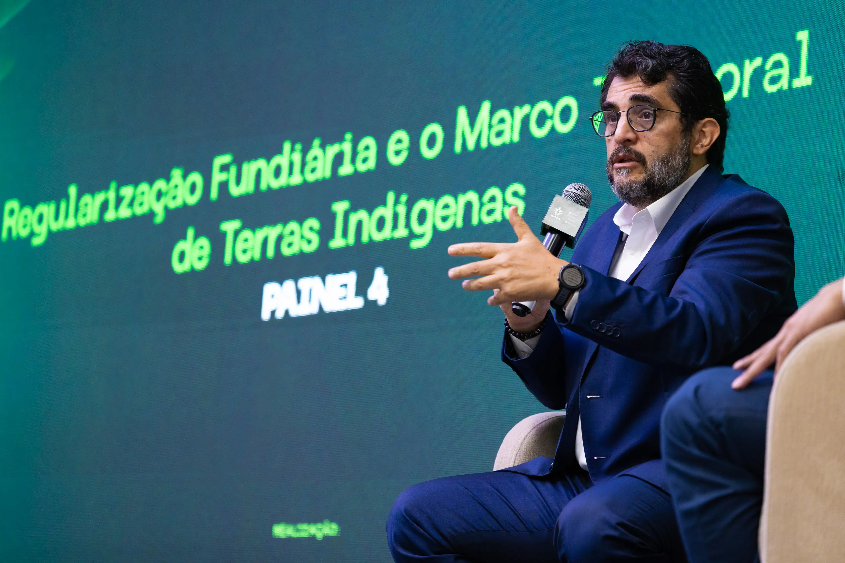 Um homem de barba e óculos, vestindo terno azul, gesticula enquanto fala ao microfone em um evento. Ao fundo, um telão verde exibe o texto sobre 