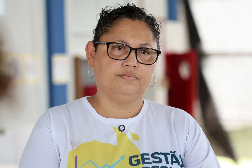 Foto horizontal em plano médio que mostra a coordenadora escolar, Amanda Lwiggy. Ela é uma mulher parda, de olhos e cabelos castanhos, usando camiseta branca com estampa da escola e óculos de grau.