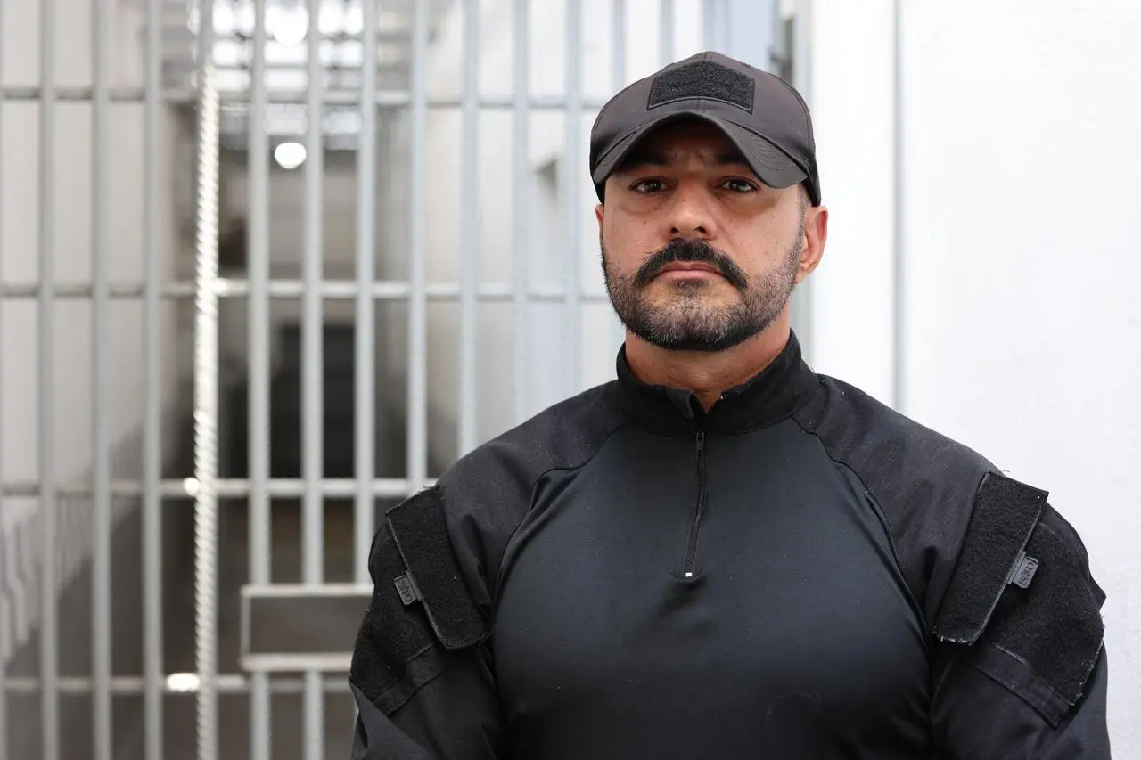 Homem de pele clara, barba e boné pretos, vestindo uniforme operacional preto. Ele encara a câmera com expressão séria. Ao fundo, uma grade de metal branca indica um ambiente prisional.