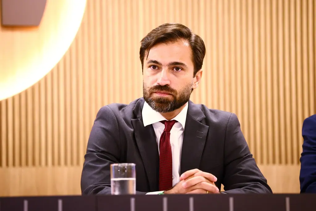 Foto horizontal em plano médio que mostra o juiz Marcelo Resende sentado no palco do auditório do TCE-MT. Ele é um homem branco, de olhos, barba e cabelos castanhos, usando camisa branca, terno cinza escuro, gravata vermelha.