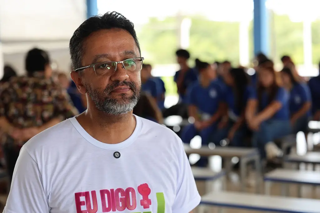 Foto horizontal em plano médio que mostra o assessor da Cemulher Cristian Pereira no pátio da escola. Ele é um homem negro, de cabelos castanhos, barba grisalha, usando óculos de grau e camiseta branca da campanha Eu Digo Basta.