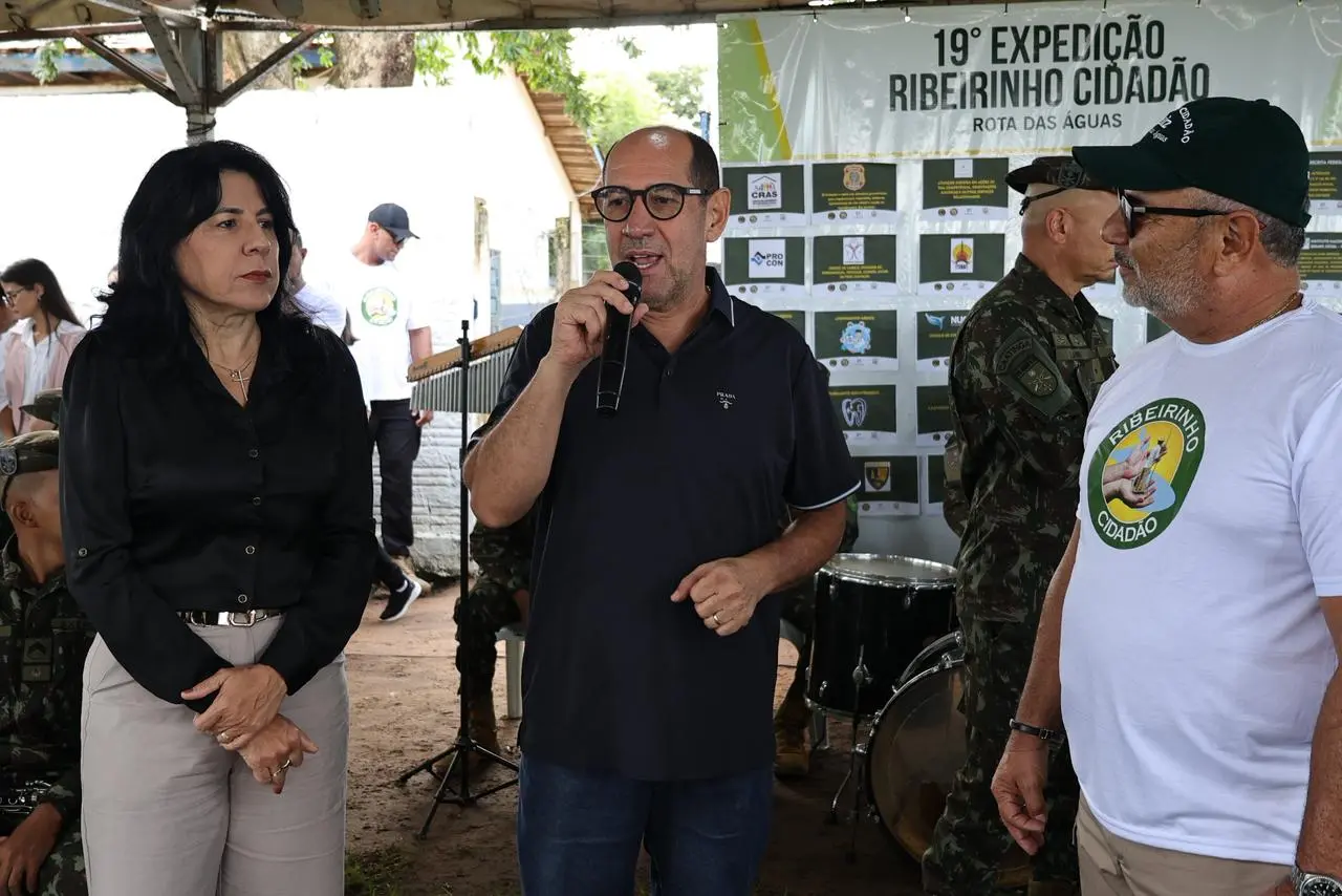 Homem de óculos e camisa polo azul fala ao microfone. Ele está entre uma mulher de blusa preta e um homem com camiseta do evento 