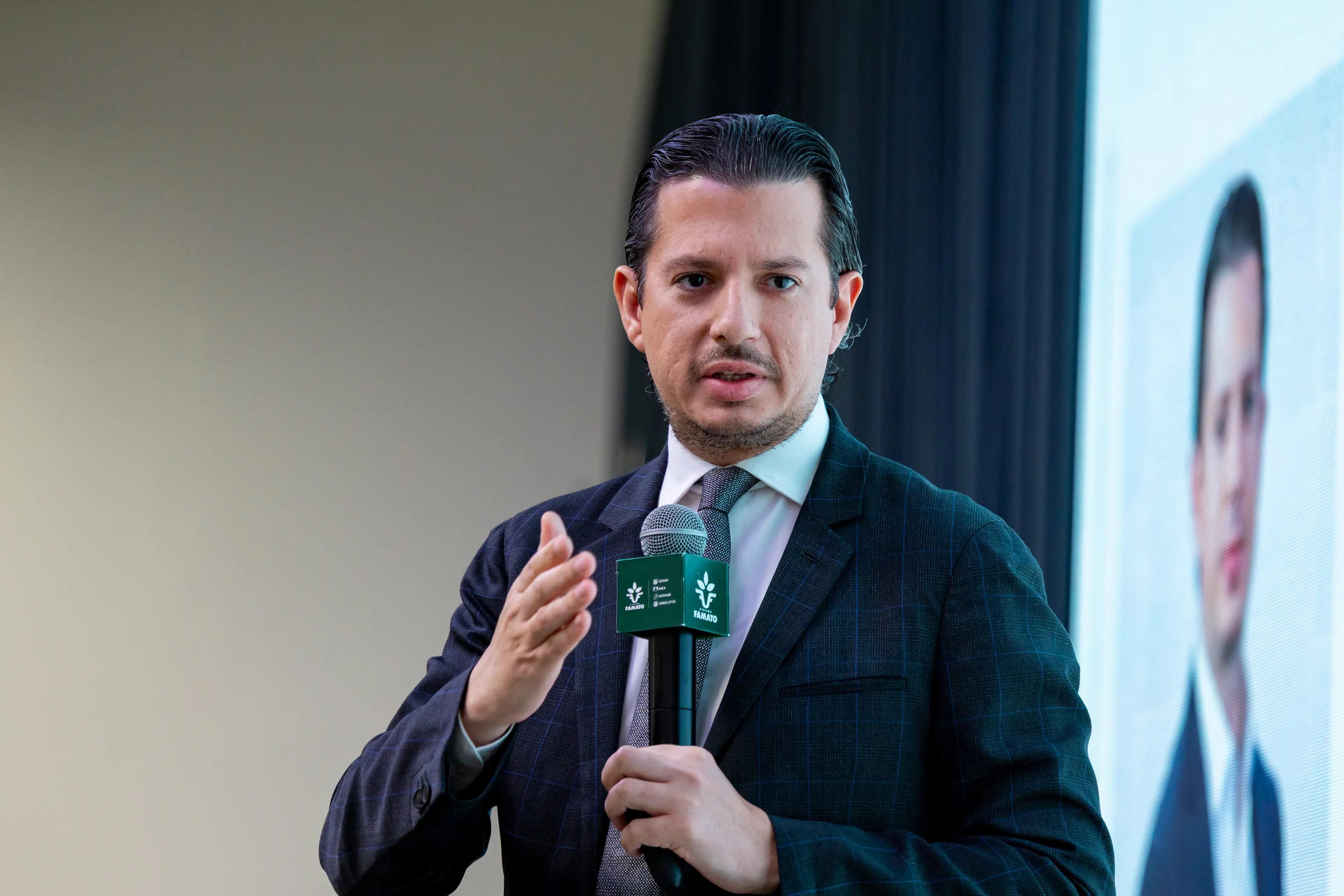 Um homem de terno escuro e gravata cinza fala ao microfone em um evento. Ele tem cabelos curtos e gesticula com a mão direita. Ao fundo, parte de um telão exibe uma imagem masculina desfocada.