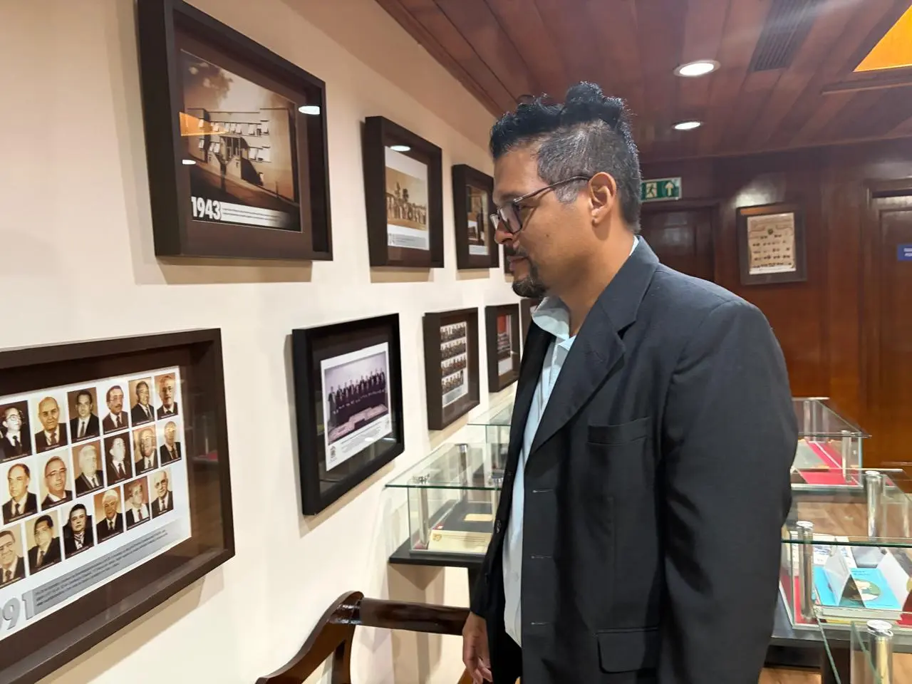 Um homem de cabelos escuros com topete, veste blazer escuro, observa atentamente fotografias históricas emolduradas na parede. Ao fundo, vitrines com acervo e mais quadros