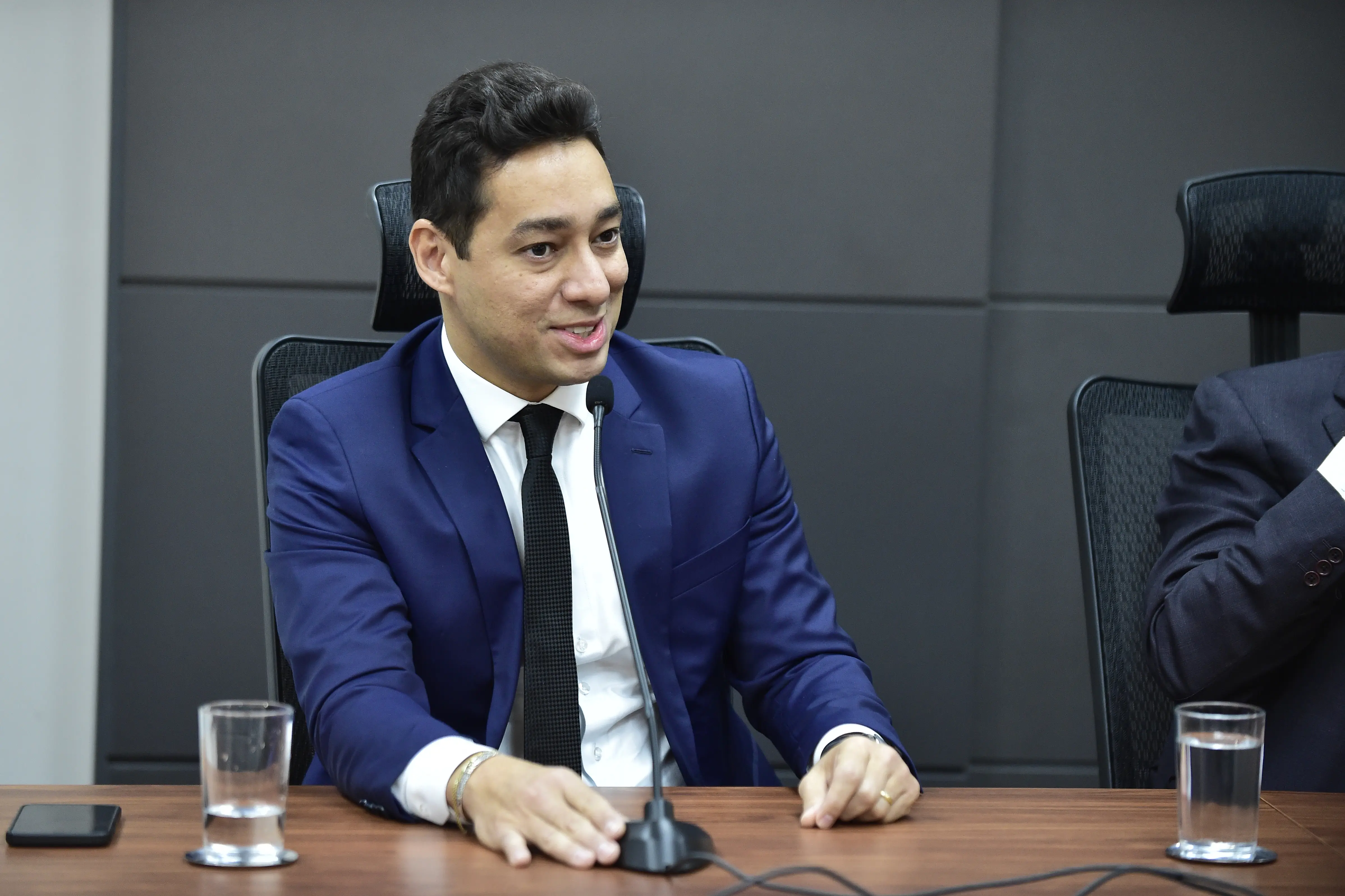 Foto que mostra o procurador-geral do Município de Cuiabá, Luiz Antônio Araújo Júnior, sentado e falando ao microfone. Ele é um homem pardo, de olhos e cabelos castanhos escuros, usando camisa branca, gravata preta e terno azul.