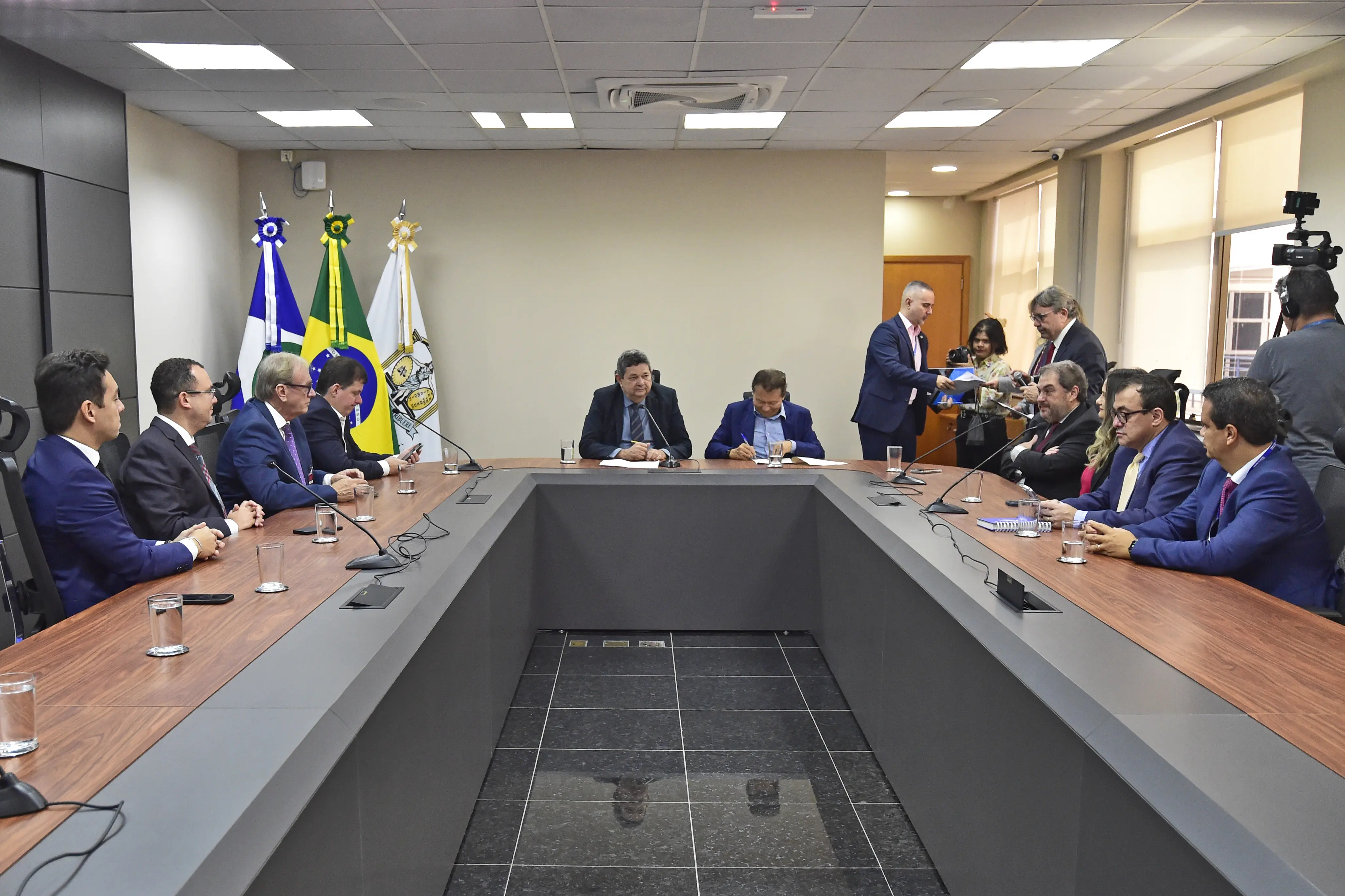 Foto em plano aberto que mostra a sala de reuniões do TJMT com desembargadores, juízes e procuradores do Estado e do Município de Cuiabá sentados em volta da mesa. No canto esquerdo, há mastros com as bandeiras do Brasil, de Mato Grosso e do Poder Judiciário. 