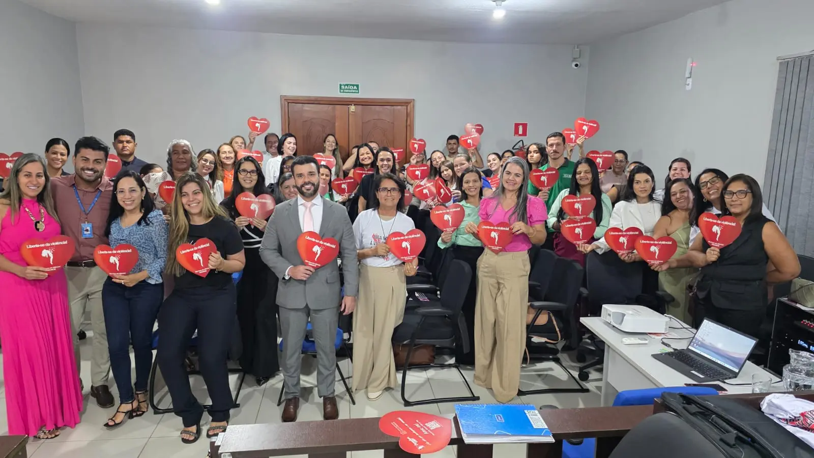 Foto horizontal que mostra quase 50 pessoas que posam sorrindo para a foto, no plenário do Fórum de Vila Rica. As pessoas estão em pé e seguram panfletos vermelhos em formato de coração.
